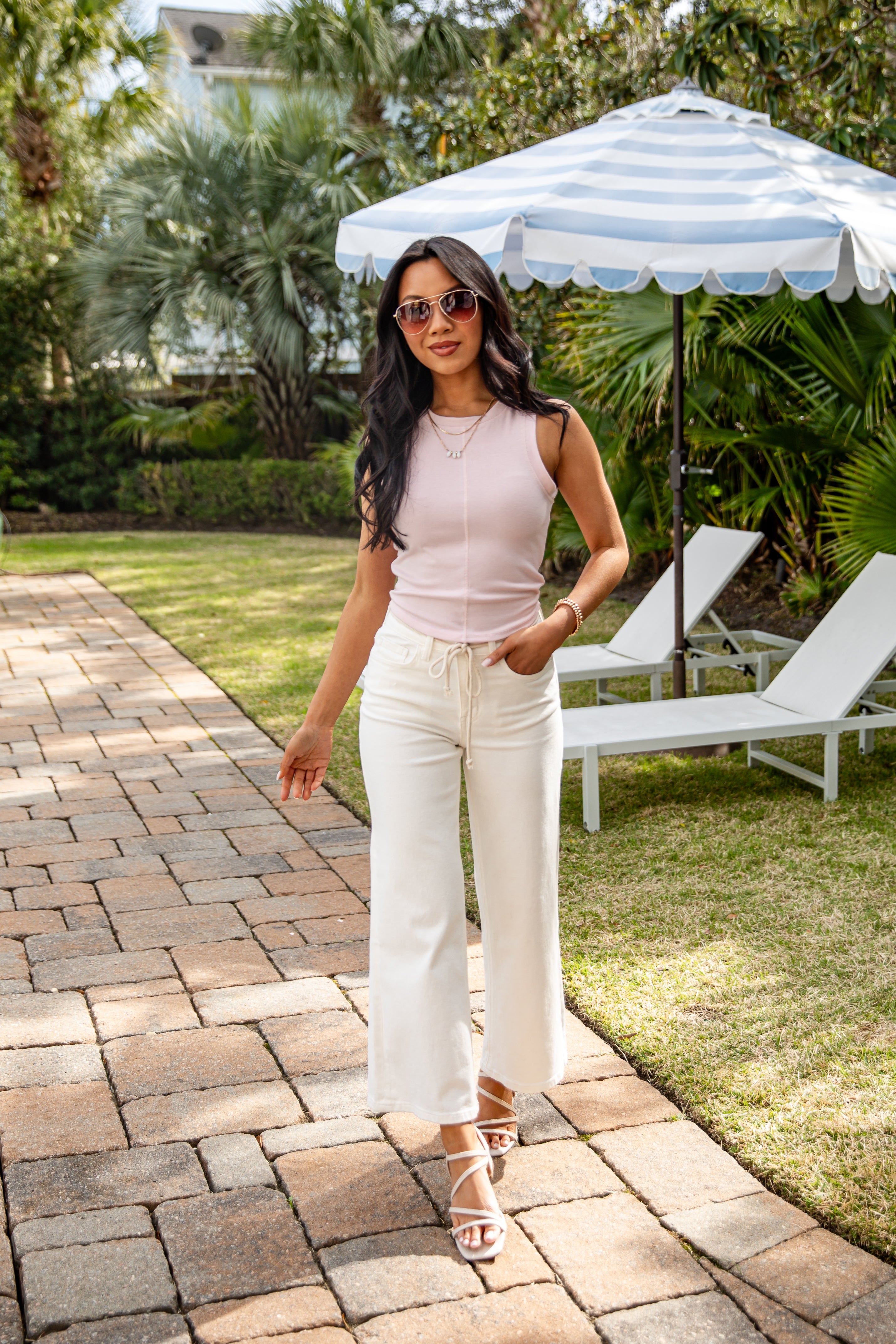 Woman in a pink top and white pants standing on a patio with lounge chairs and an umbrella in the background.
