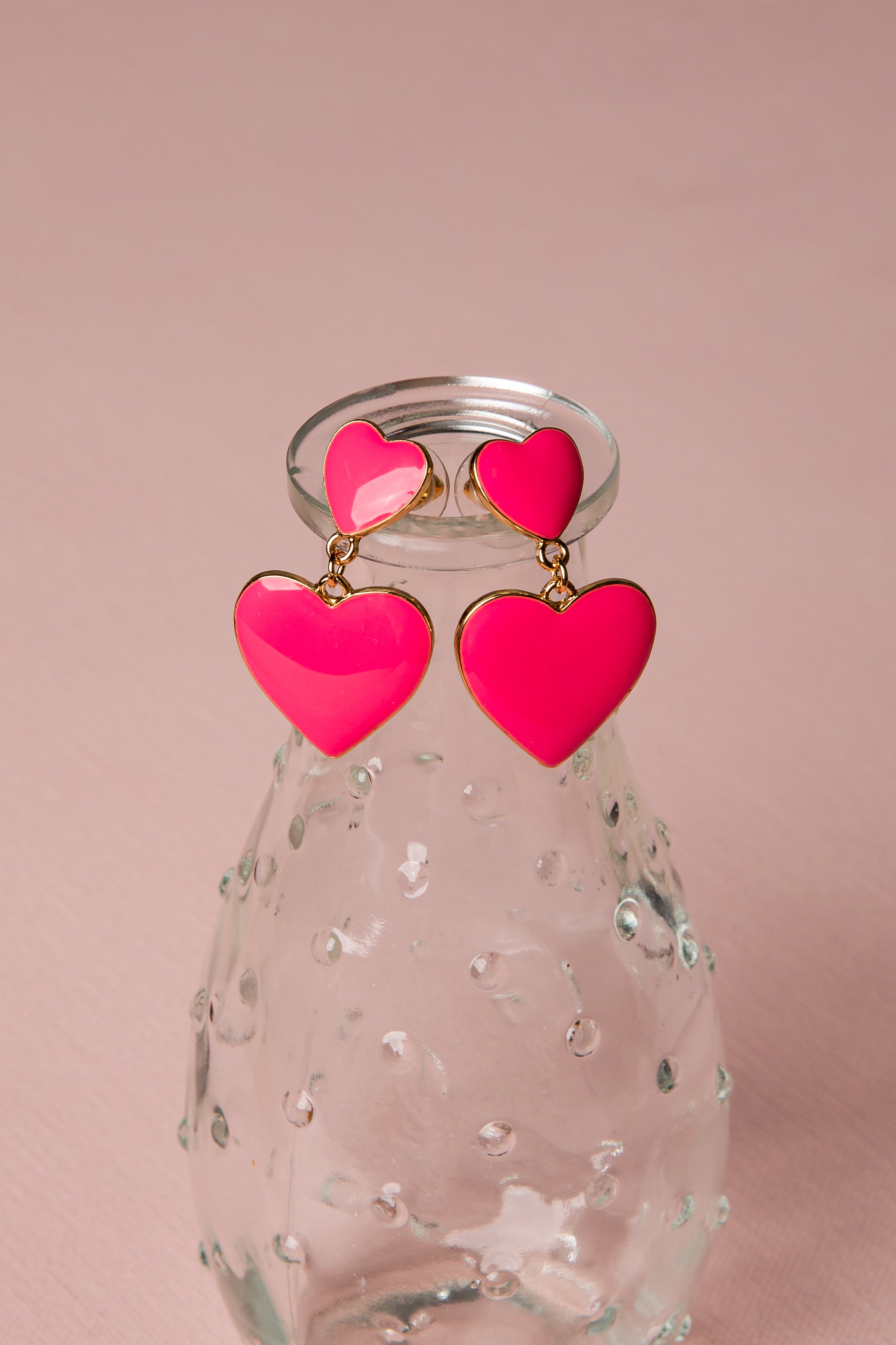 Pink heart-shaped earrings on a clear glass bottle with a pink background