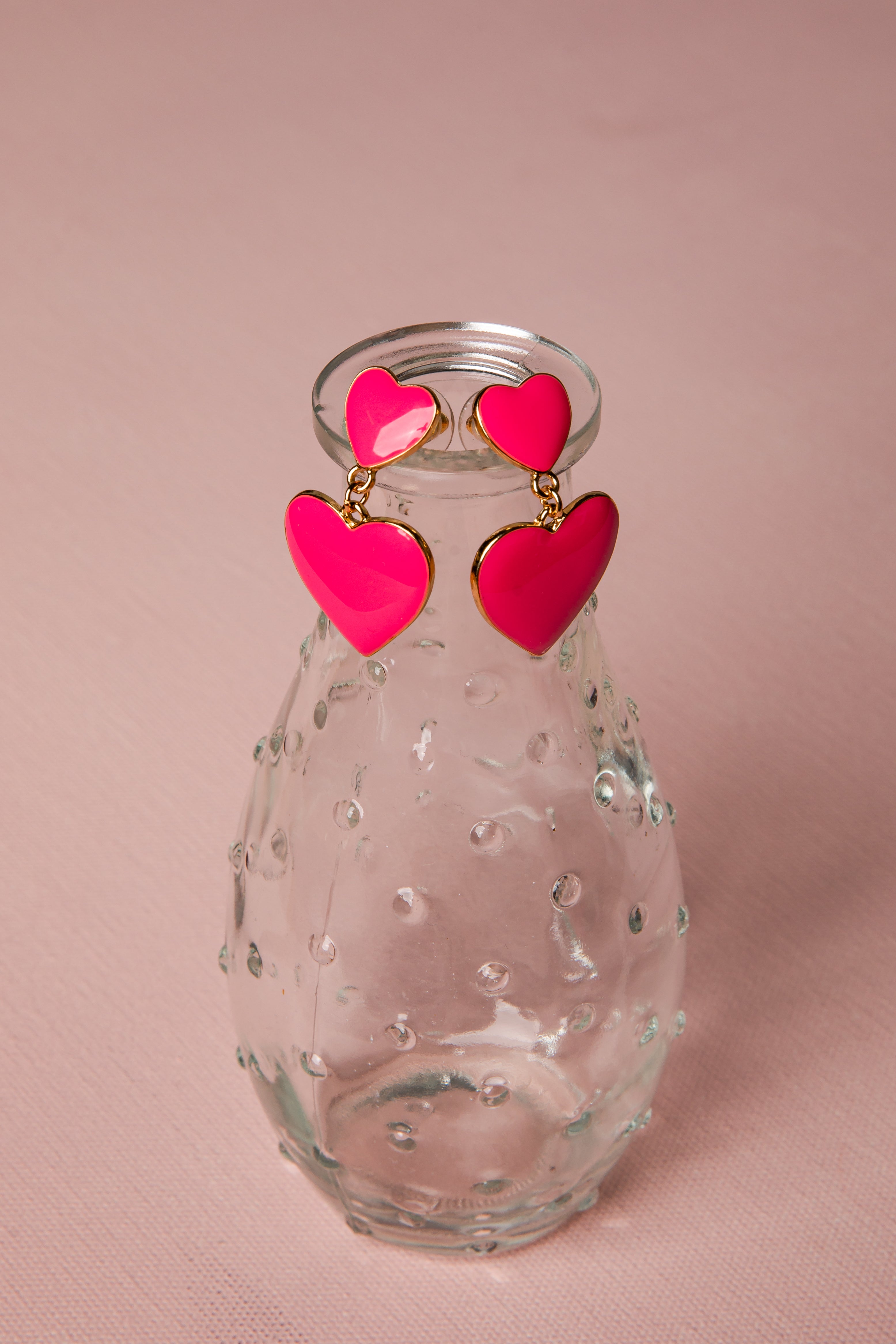 Clear glass vase with pink heart-shaped earrings on a pink background