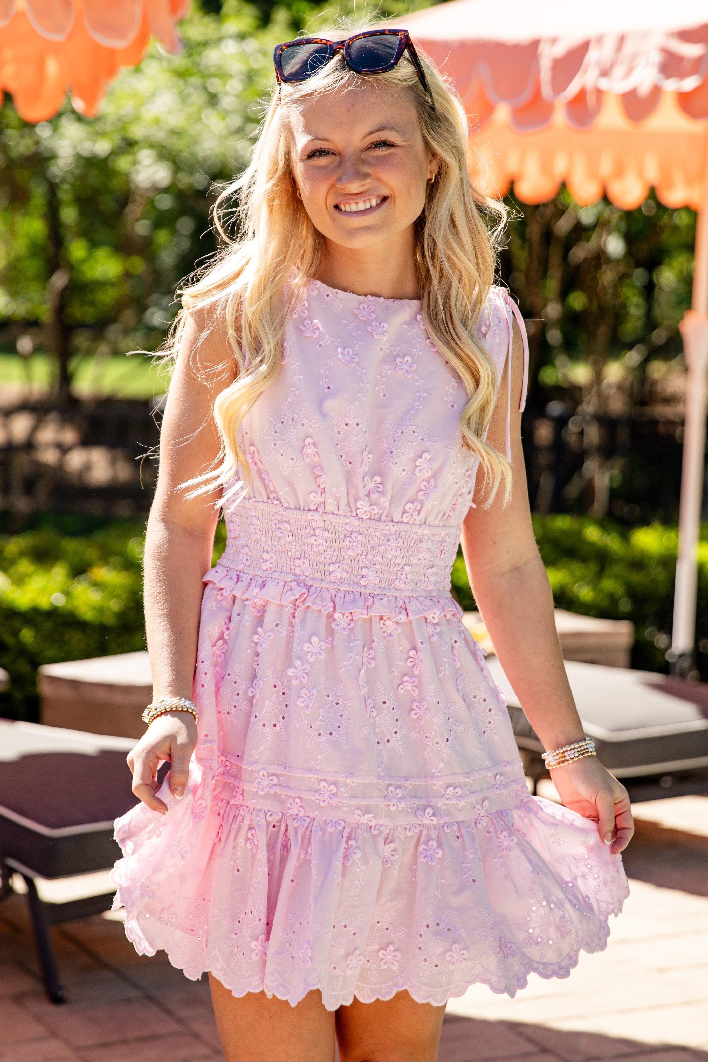 Model wearing a light pink embroidered eyelet mini dress with tie shoulder straps, a smocked waist, and a tiered ruffle skirt.