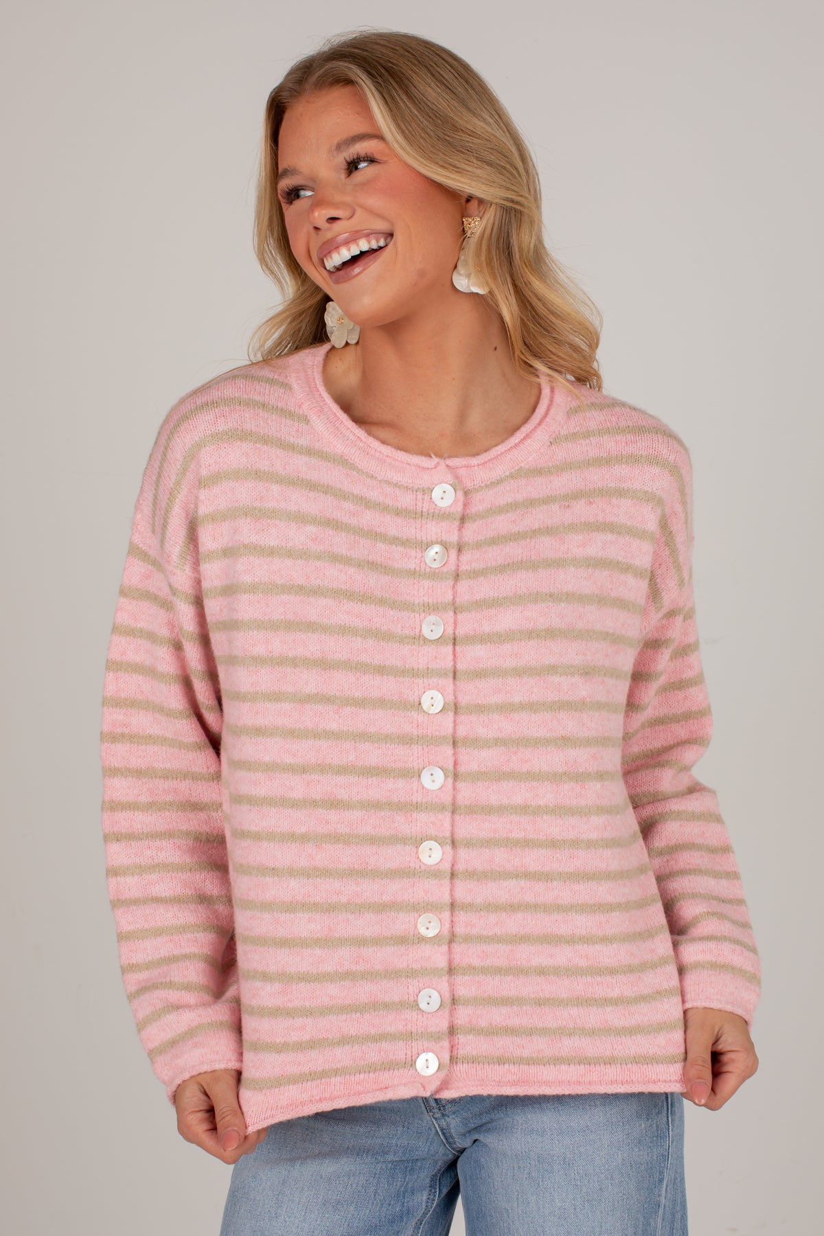 A woman wearing a pink and beige striped button-up cardigan sweater with white buttons, paired with light blue jeans, against a solid light gray background.