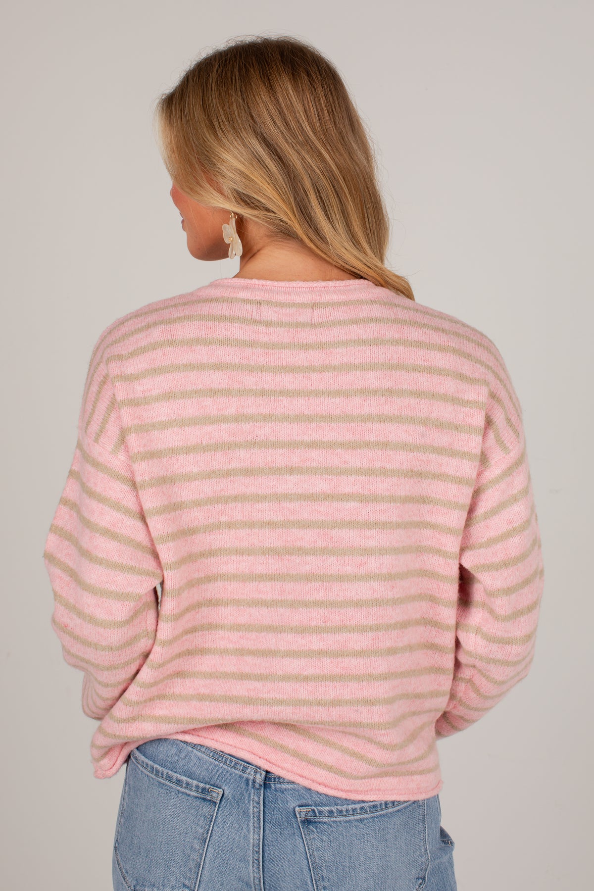 A woman viewed from the back wearing a pink and beige horizontally striped knit sweater and light blue jeans against a plain light gray studio background.