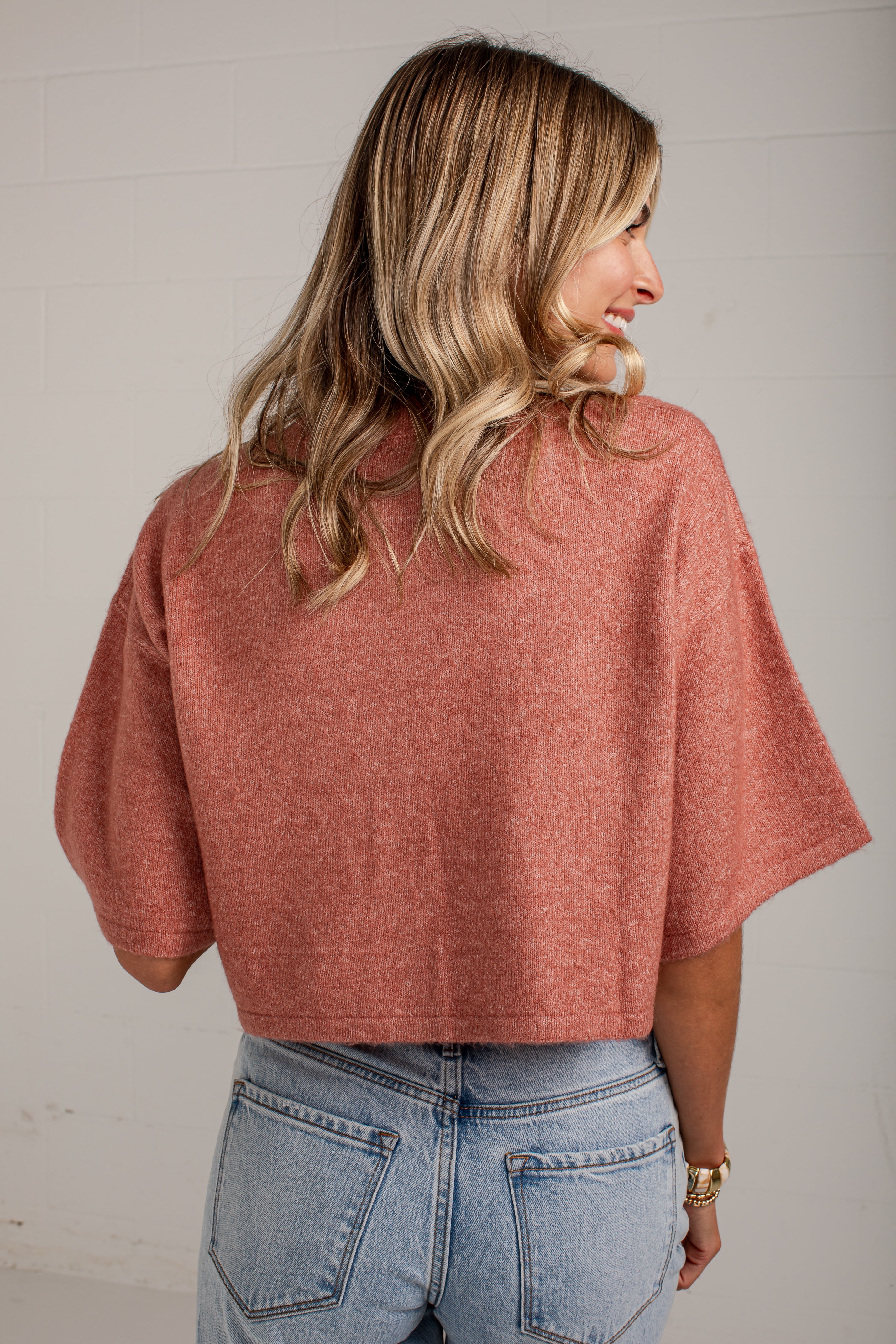 A woman shown from the back wearing a dusty rose short-sleeve knit top and light blue jeans, standing against a white paneled wall.