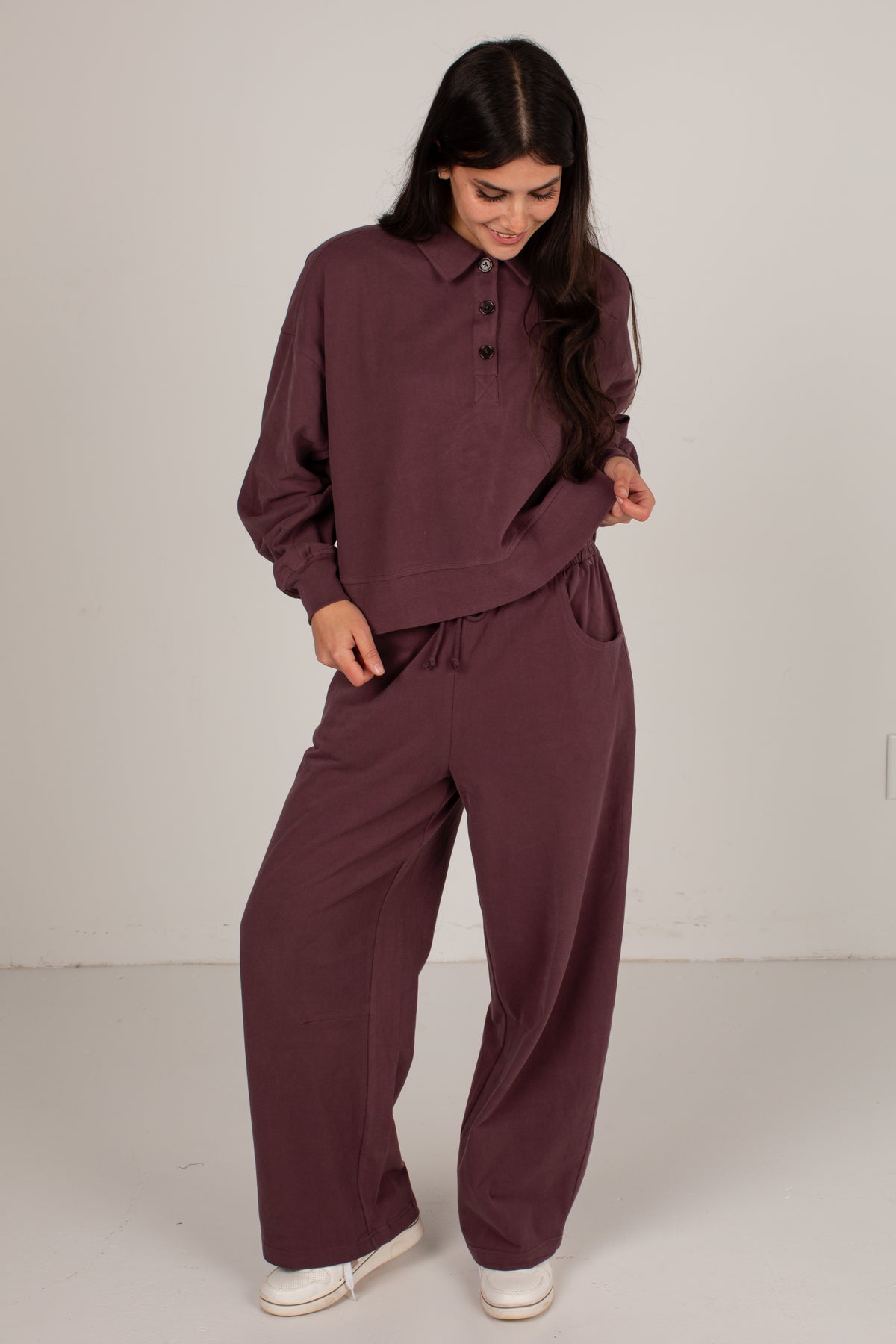 A woman modeling a matching two-piece lounge set in a deep plum purple color, featuring a long-sleeve button-up top and wide-leg pants, paired with white slip-on shoes against a plain light gray studio background.