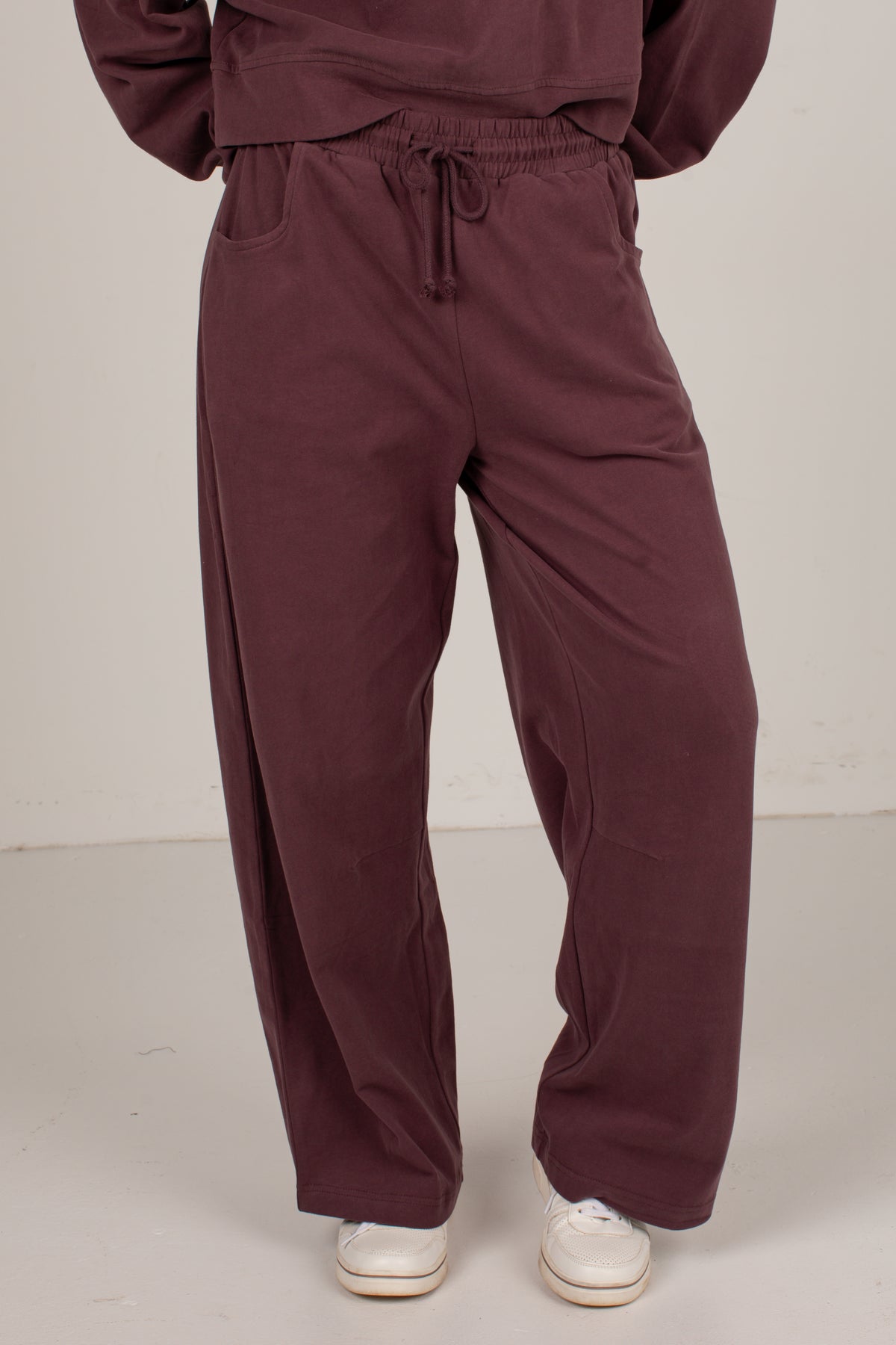 A person wearing loose, wide-leg drawstring sweatpants in a deep plum-burgundy color, paired with white sneakers, shown from the waist down against a plain off-white studio background.