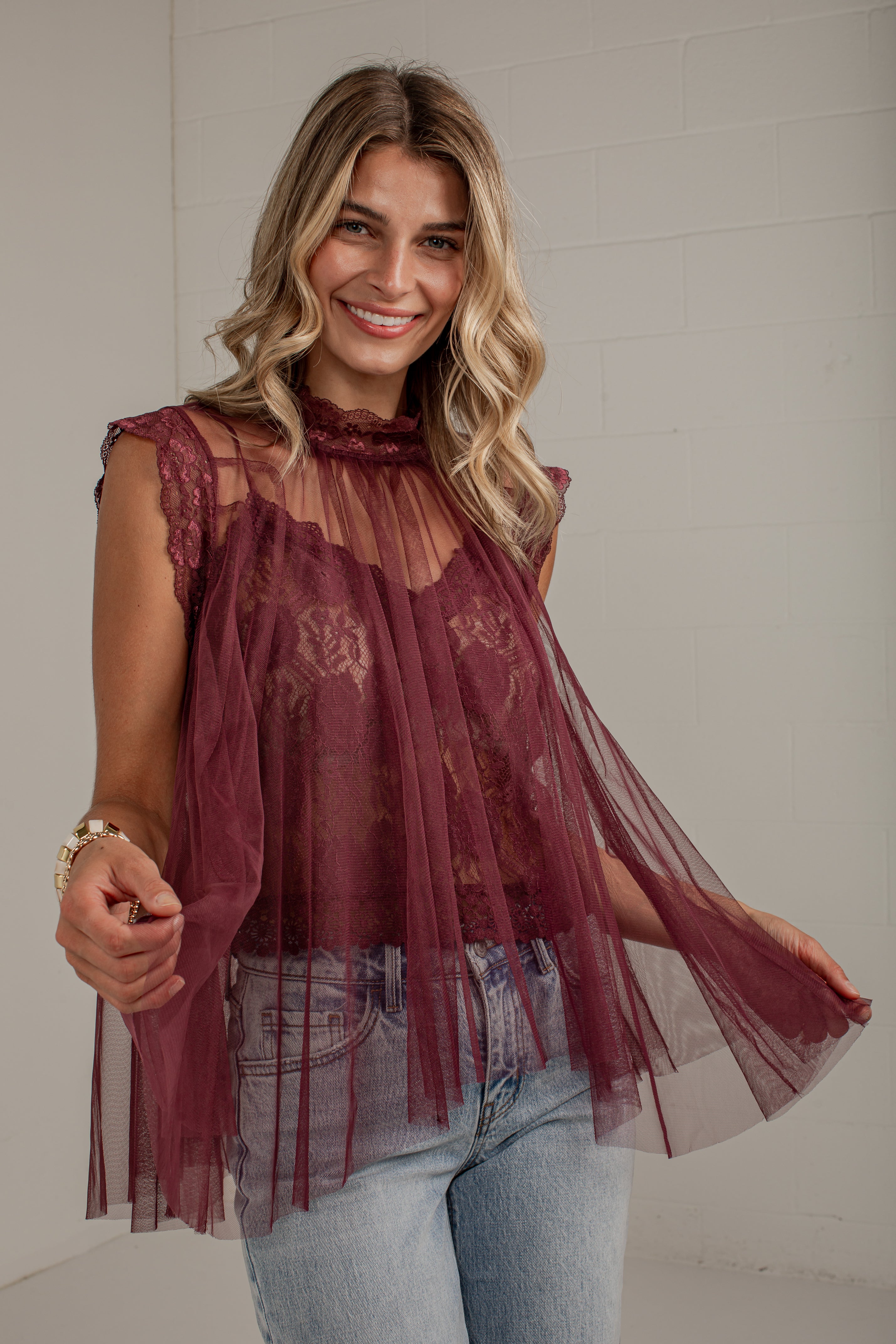 A woman wearing a sheer burgundy lace and tulle sleeveless top with light blue jeans, standing against a white brick wall.