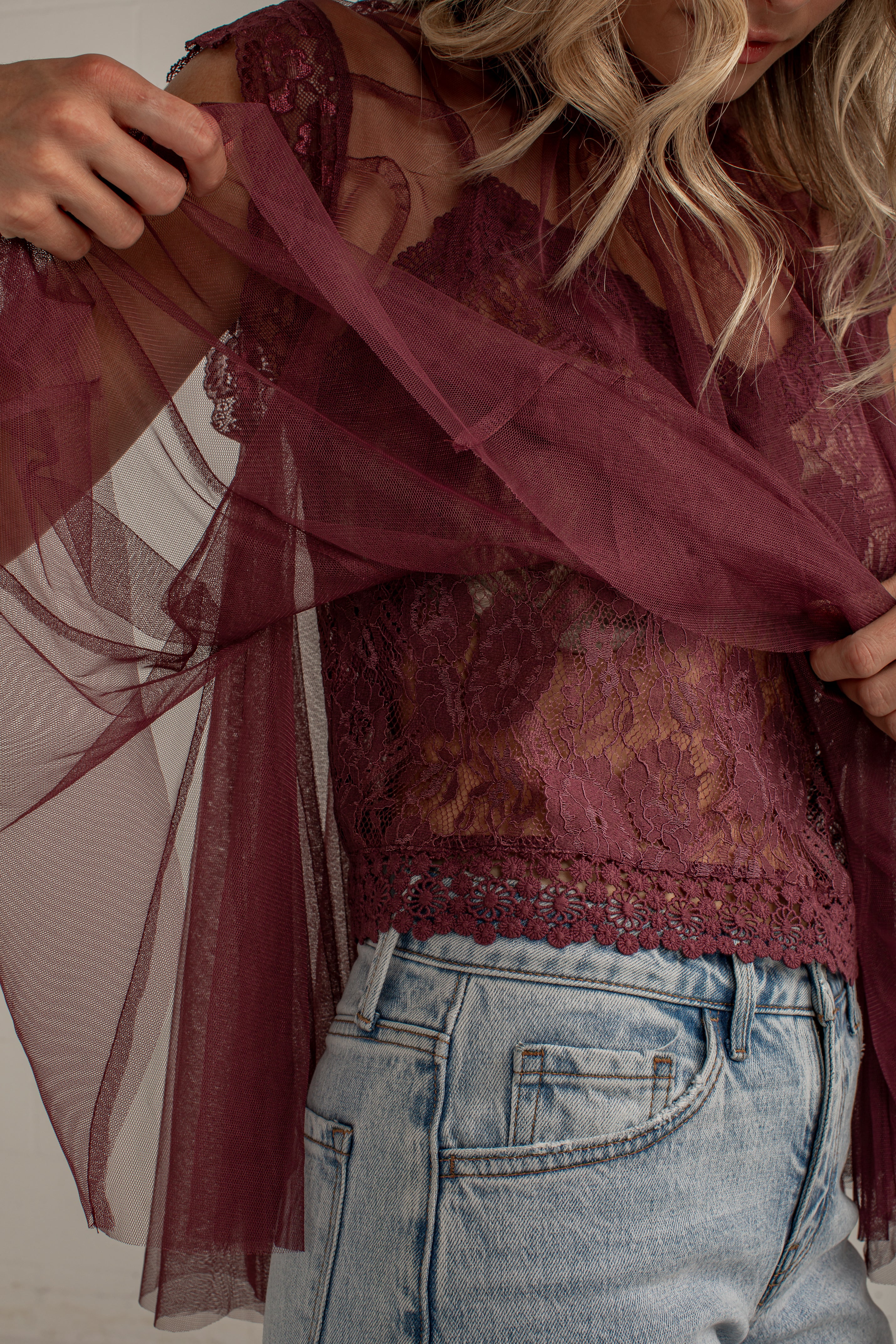 A close-up of a woman wearing a sheer burgundy tulle overlay and matching lace camisole top, paired with light blue jeans.