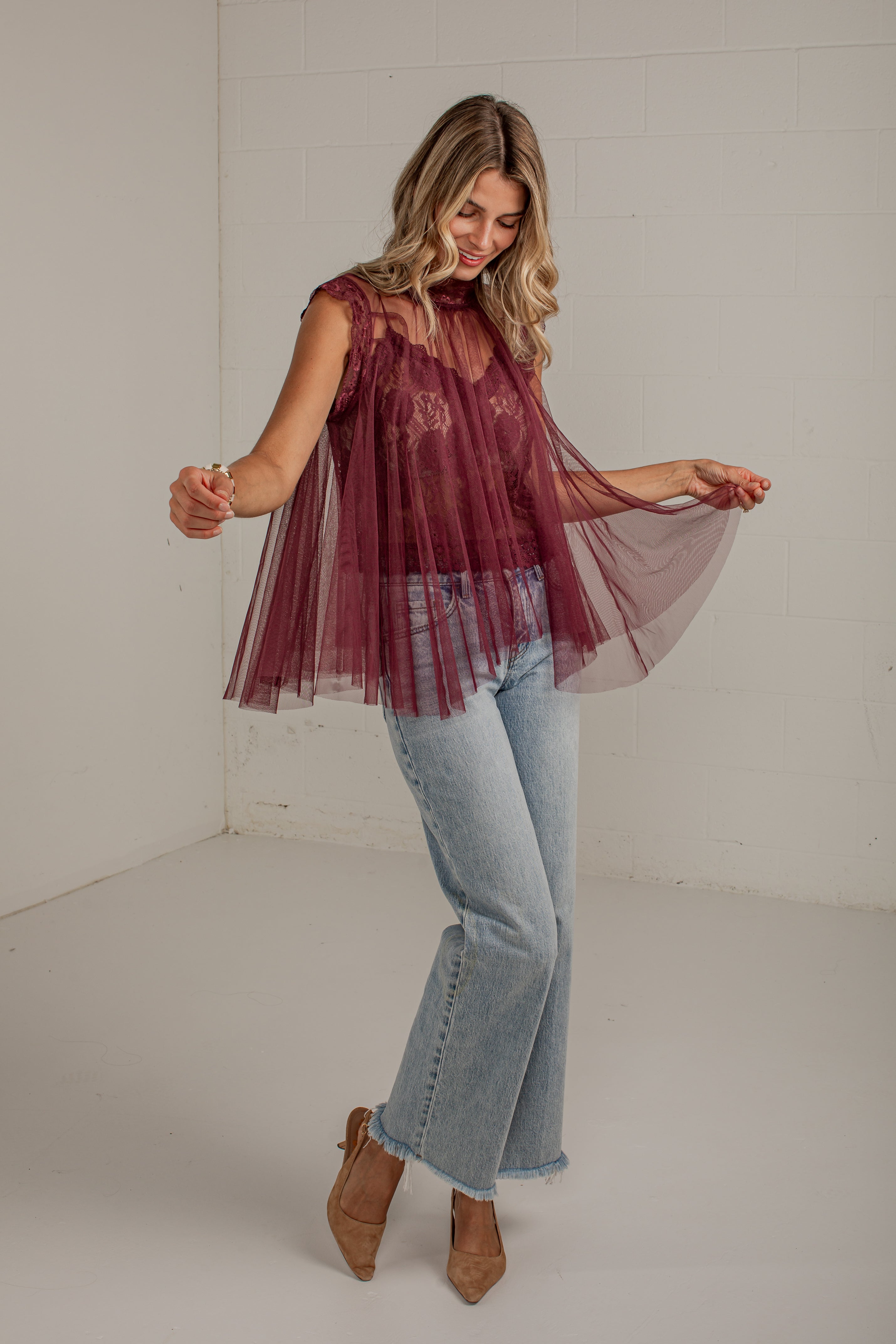 A woman modeling a sheer burgundy tulle top with a floral lace overlay, paired with light-wash flare jeans and brown ankle boots, posing in a bright white studio corner.