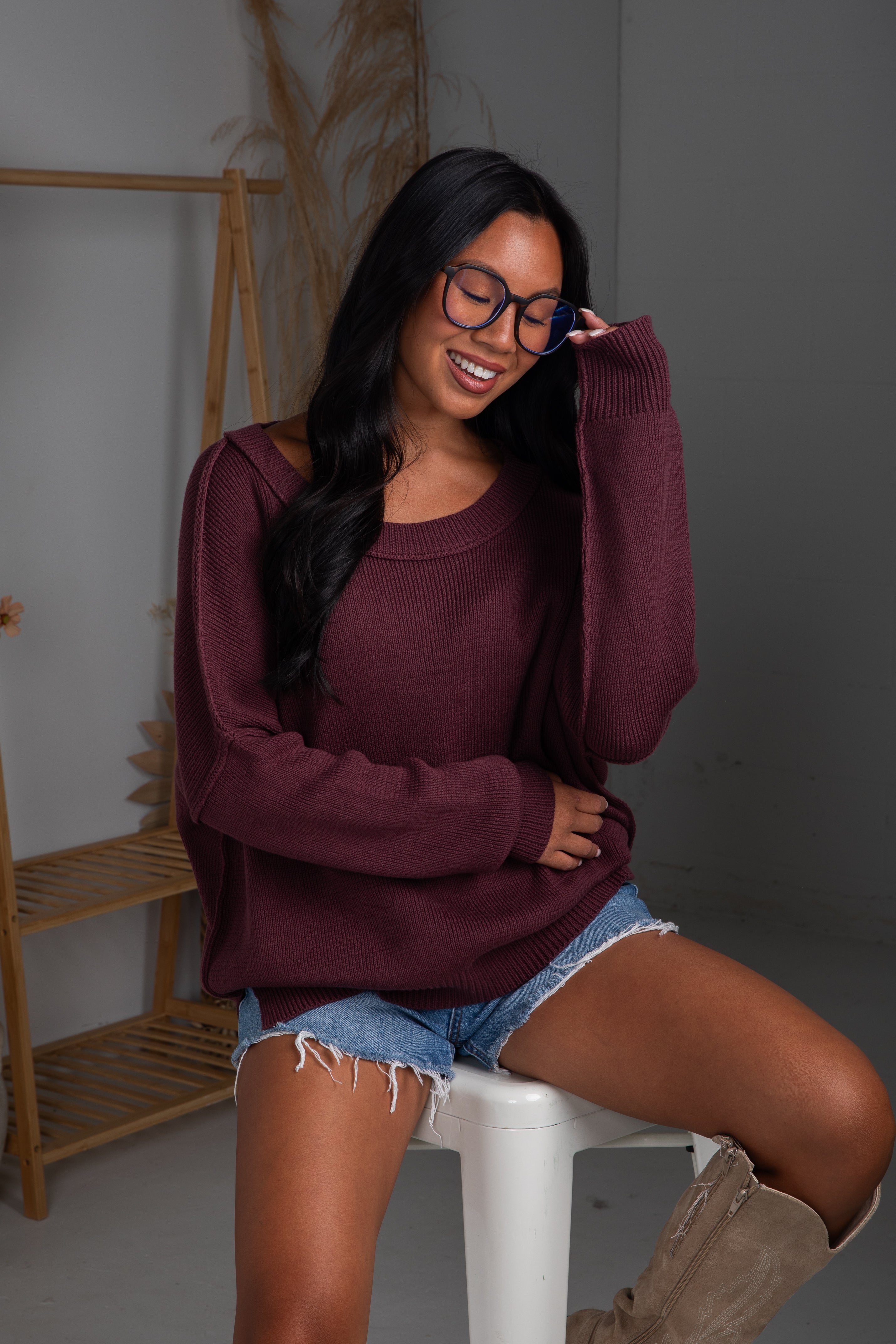 Woman wearing a maroon sweater and denim shorts sitting on a white stool.