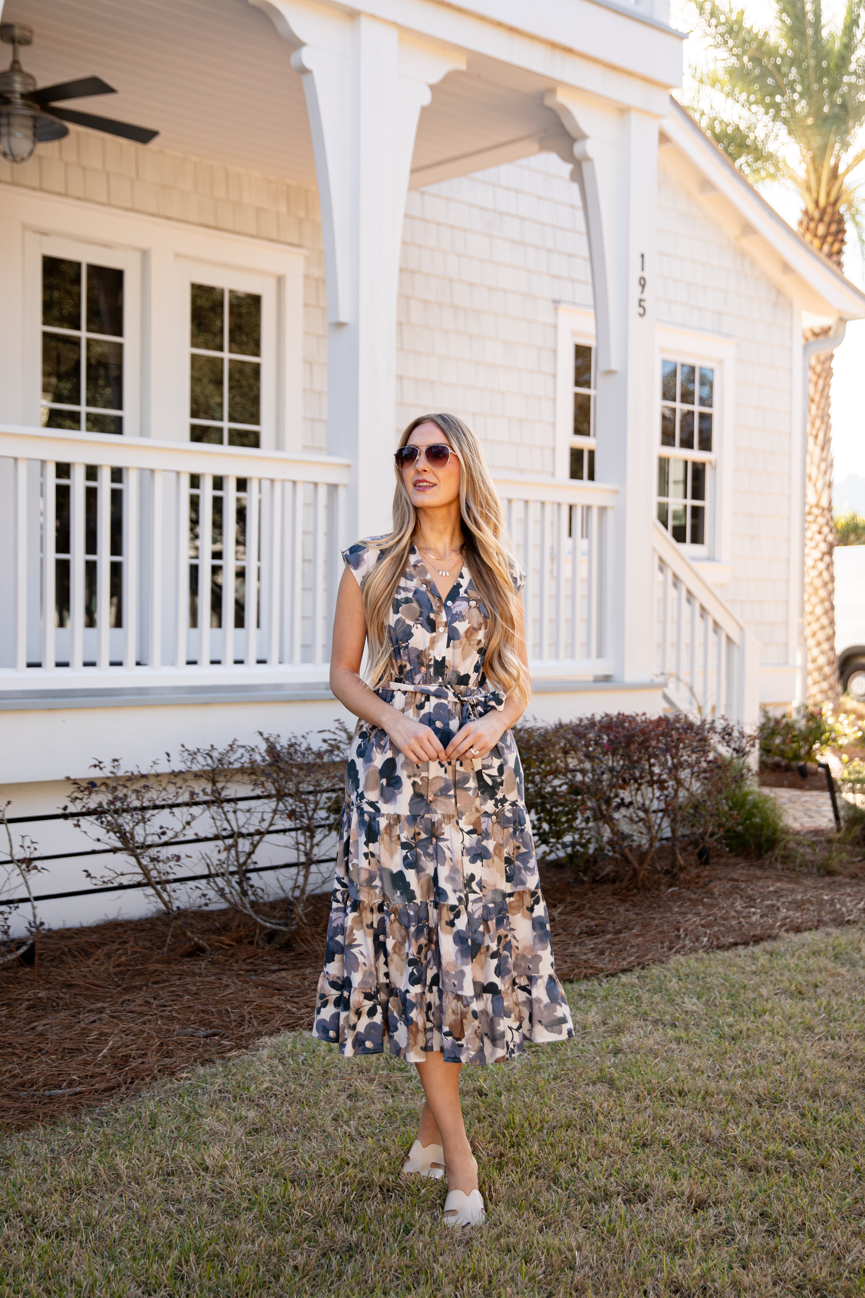 Woman in a floral dress standing on a porch with white columns and a railing.