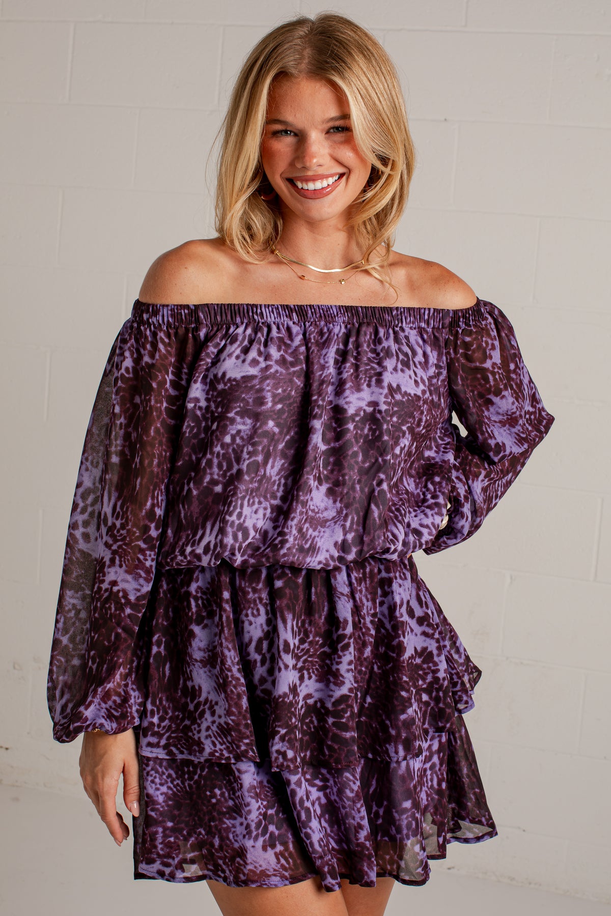 A smiling blonde woman wearing a purple and lavender tie-dye off-the-shoulder mini dress with long sheer sleeves, standing against a light gray brick wall.