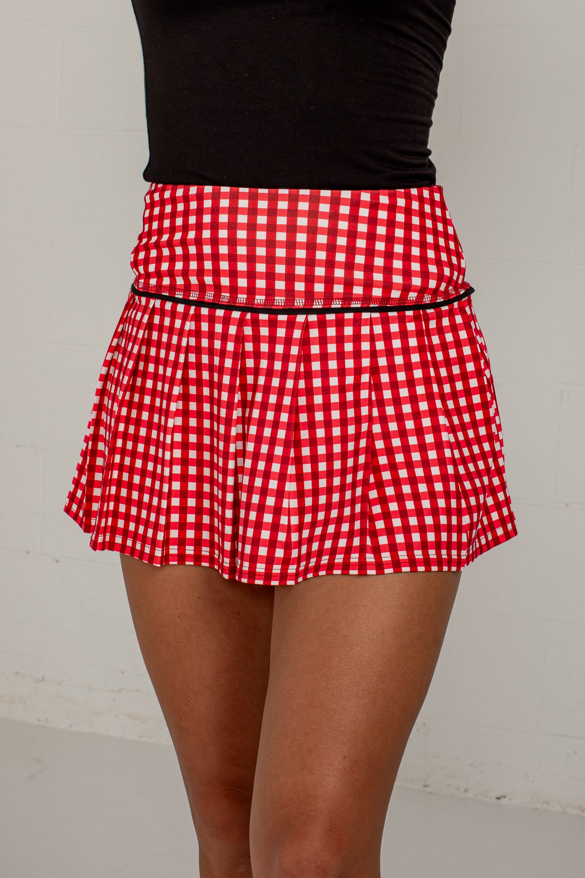A red and white gingham pleated mini skirt with a thin black waistband trim, shown on a model wearing a black top against a light gray wall.