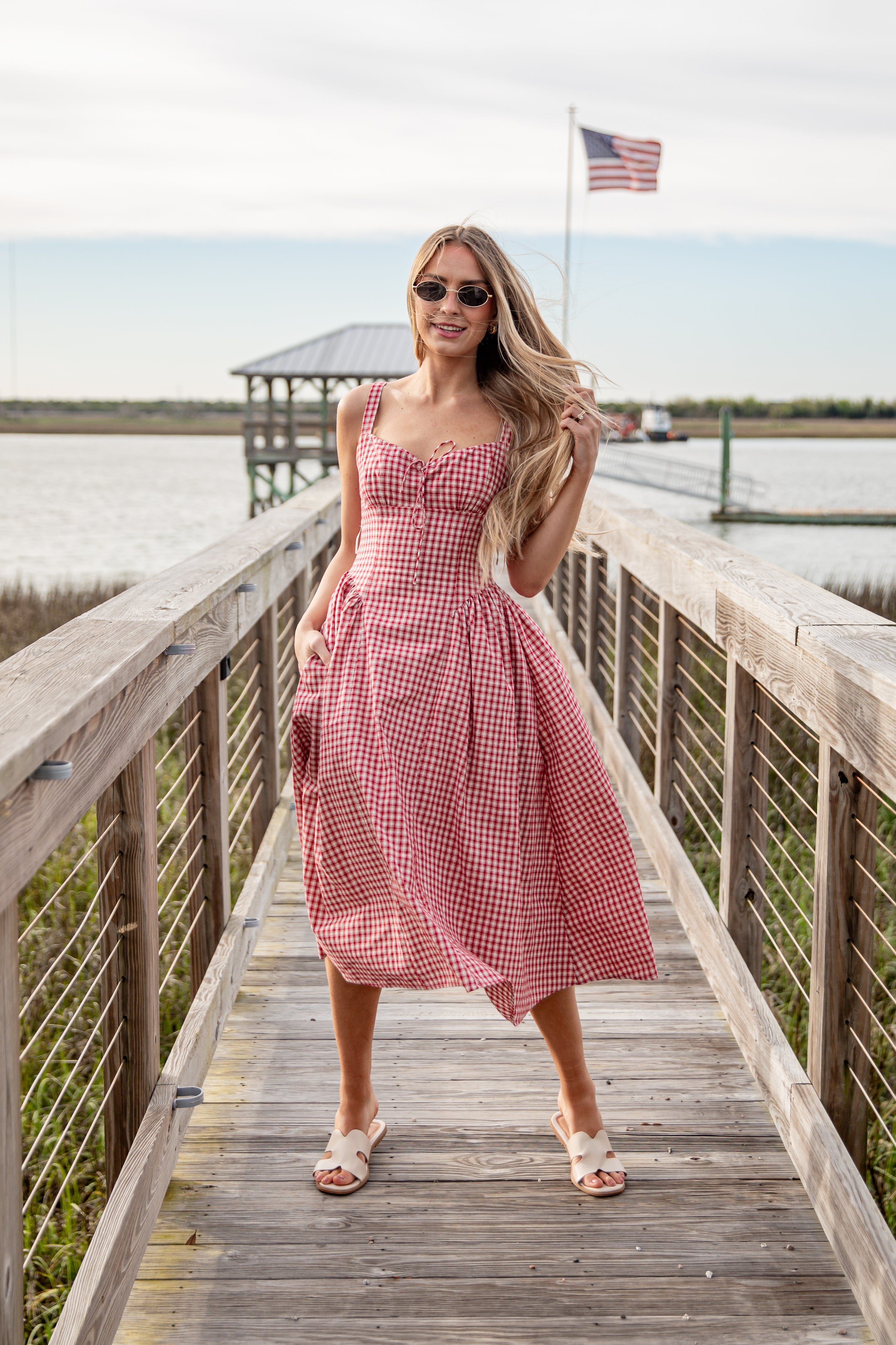 Woman in a red checkered dress standing on a wooden dock with an American flag in the background.