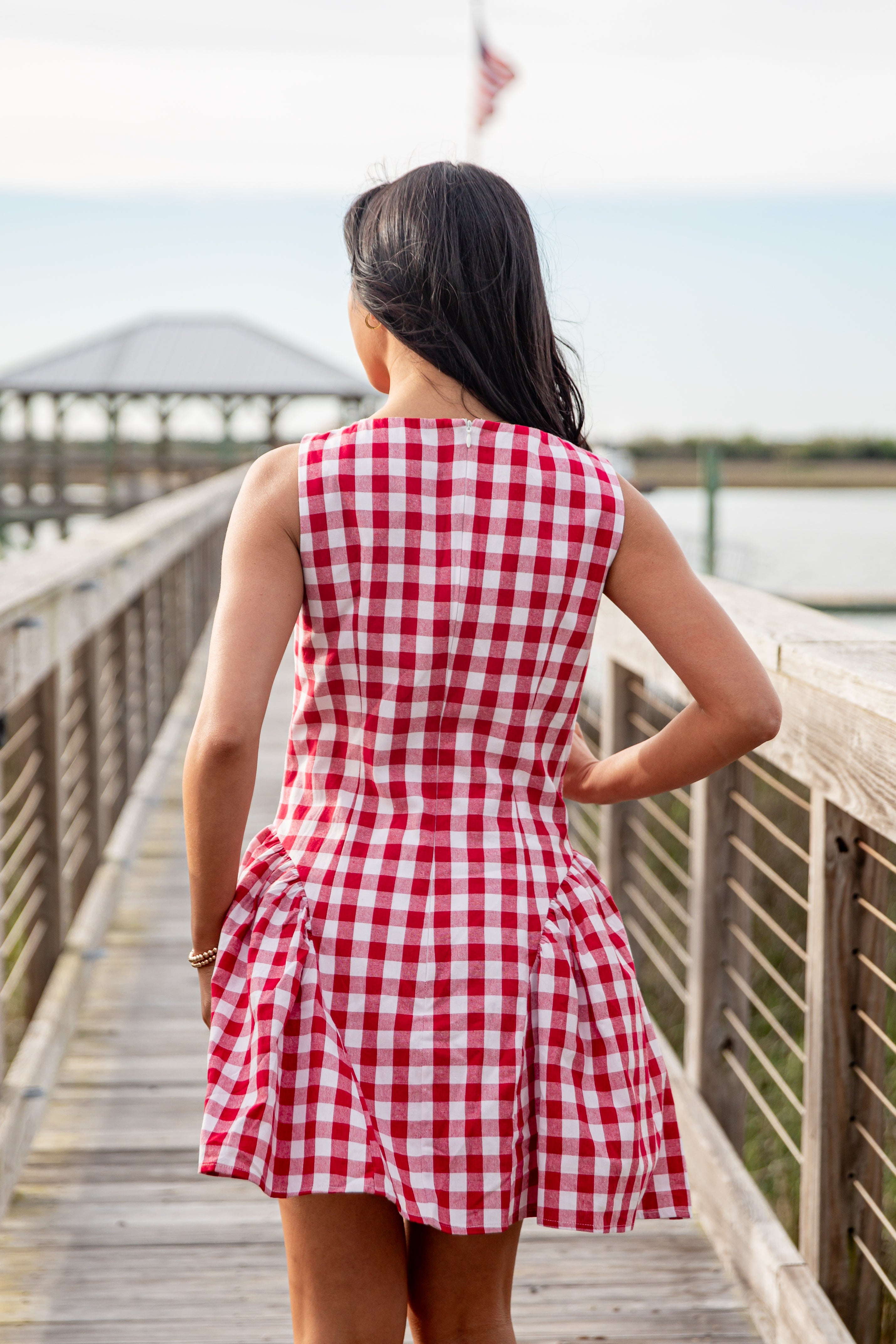 Woman in a red and white checkered dress standing on a wooden boardwalk by a waterfront.