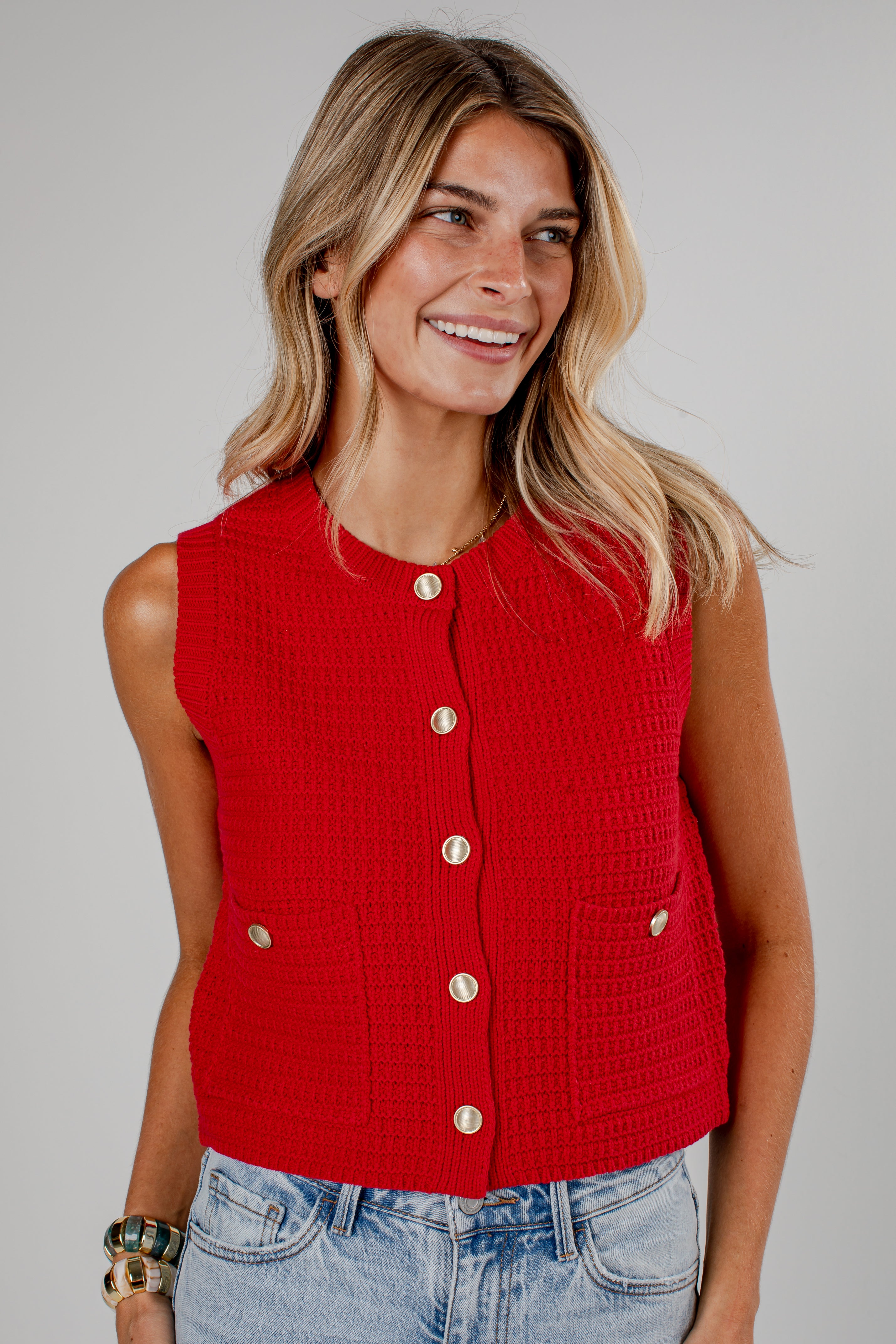 A woman wearing a bright red sleeveless cropped knit vest with gold-tone buttons and front pockets, paired with light-wash blue jeans, against a solid light gray studio background.