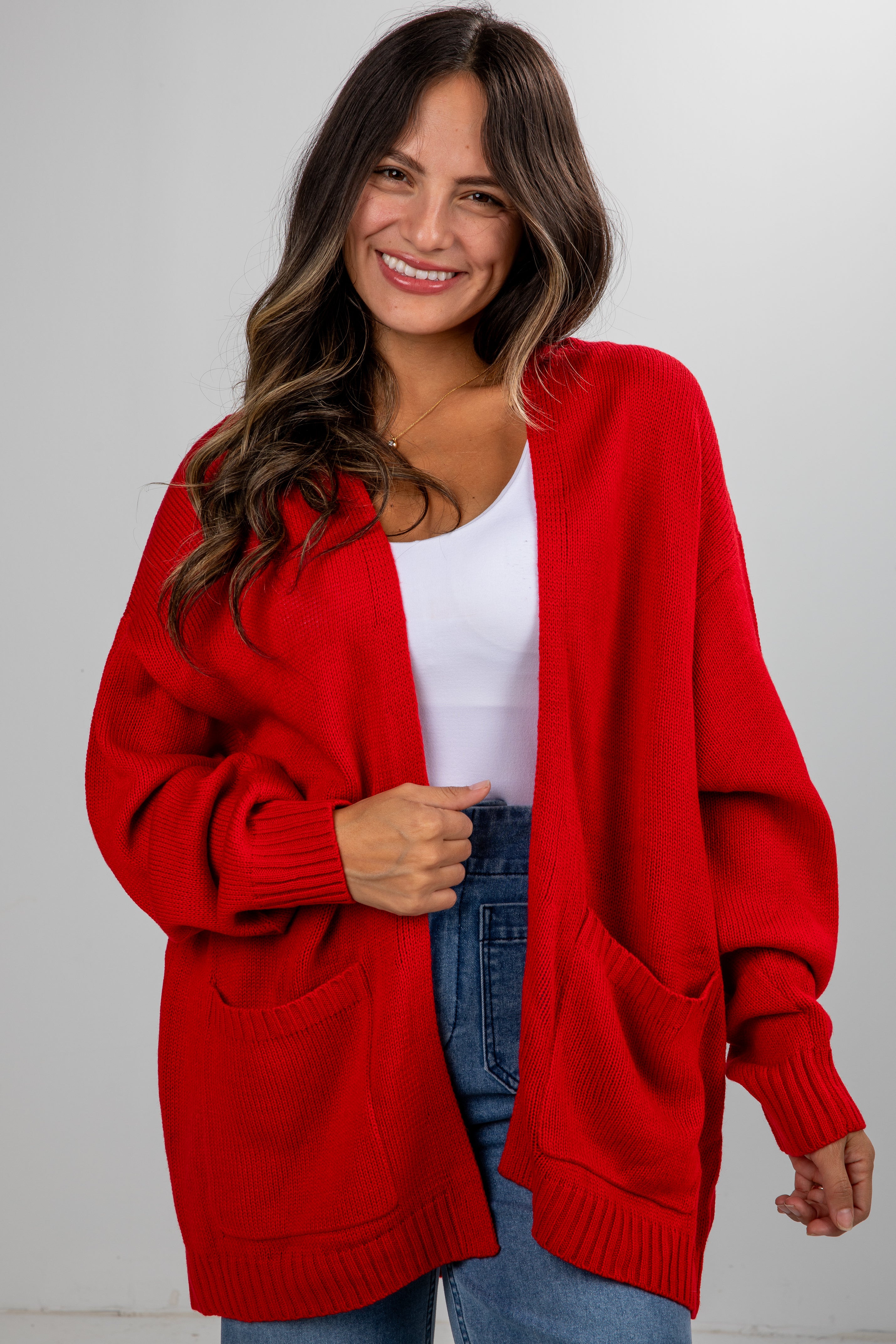 Woman wearing a red cardigan over a white shirt and blue jeans on a plain background