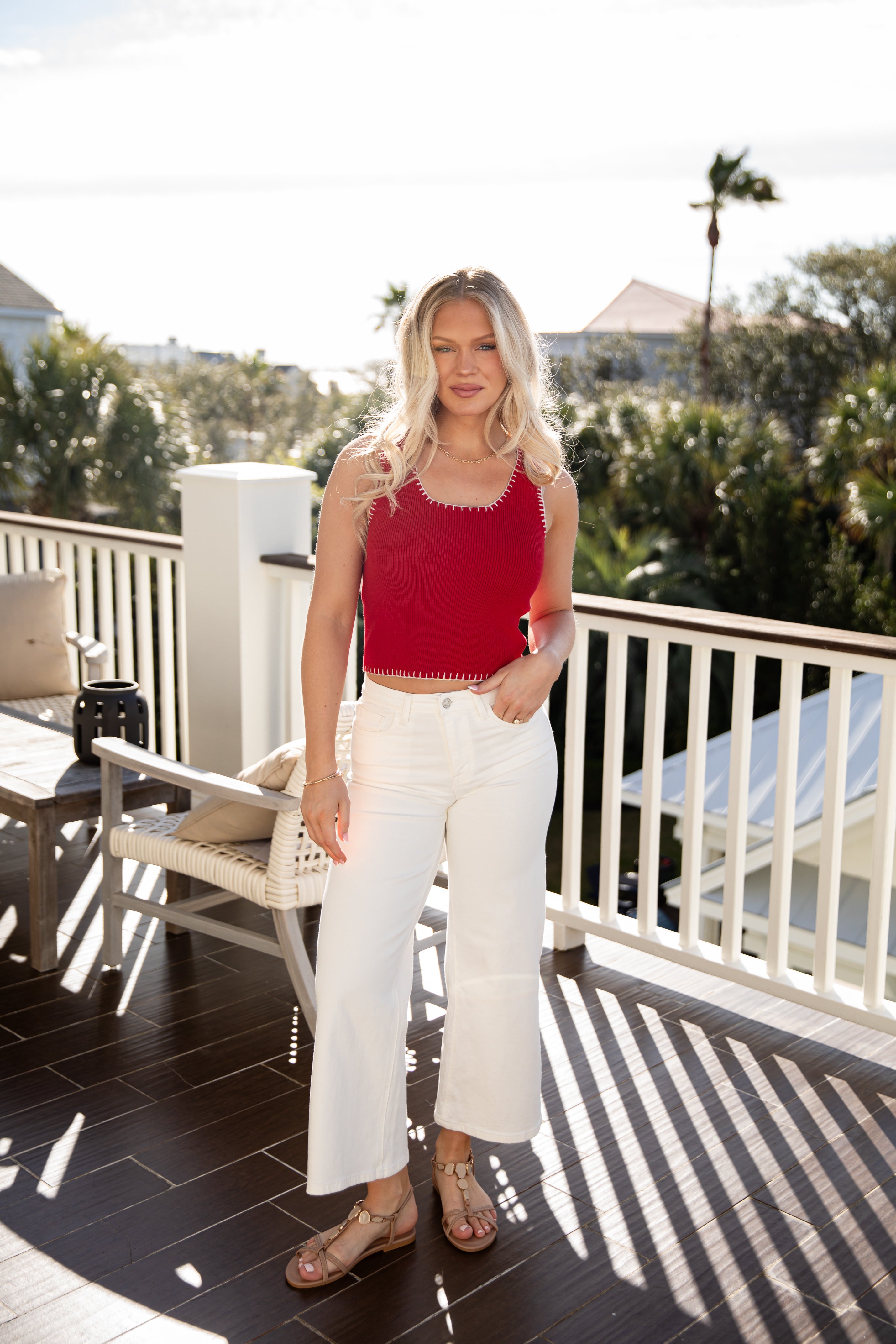 Woman in red top and white pants standing on a balcony with trees and buildings in the background