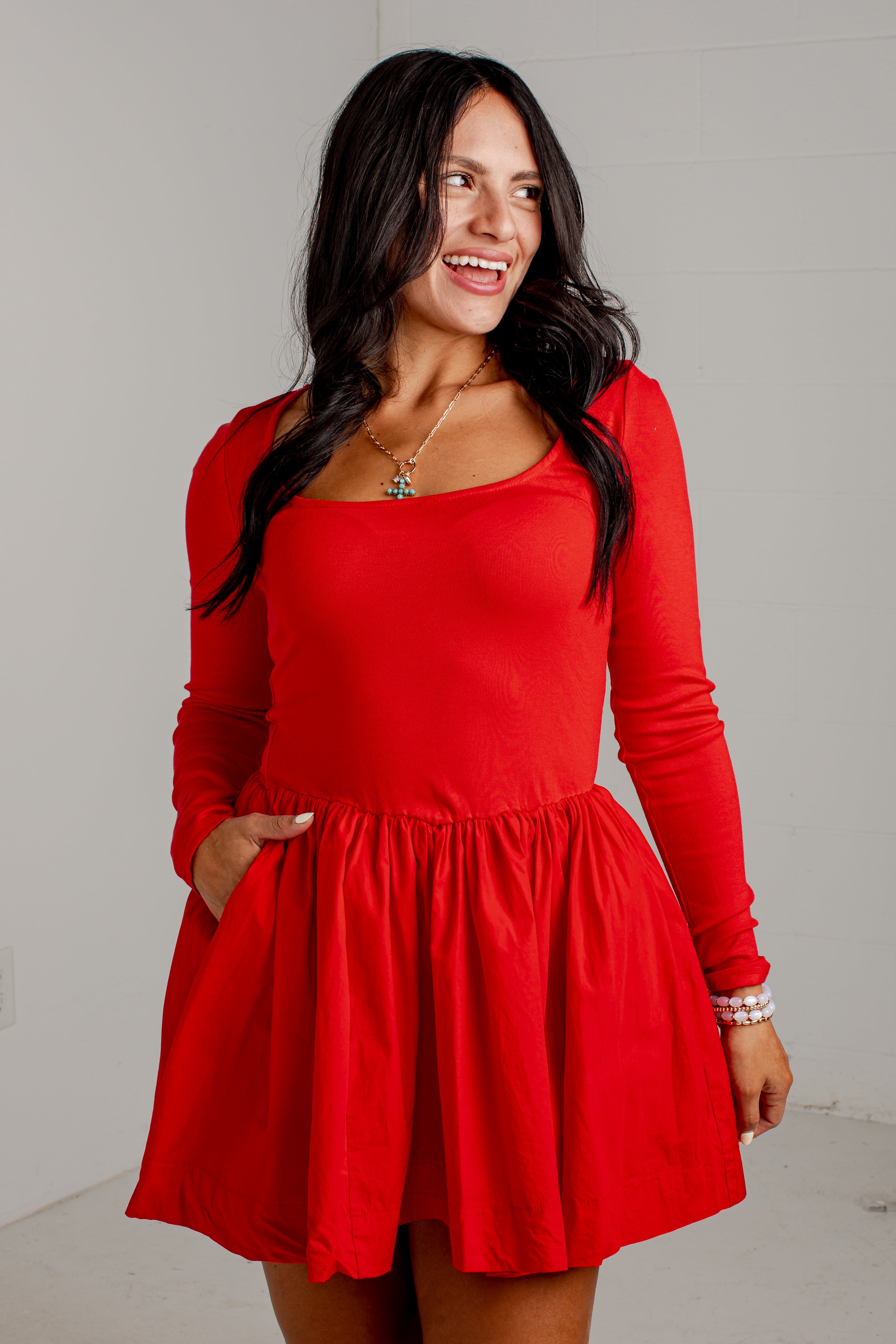 A woman modeling a bright red long-sleeve skater dress with a scoop neckline and side pockets, standing against a light gray wall.