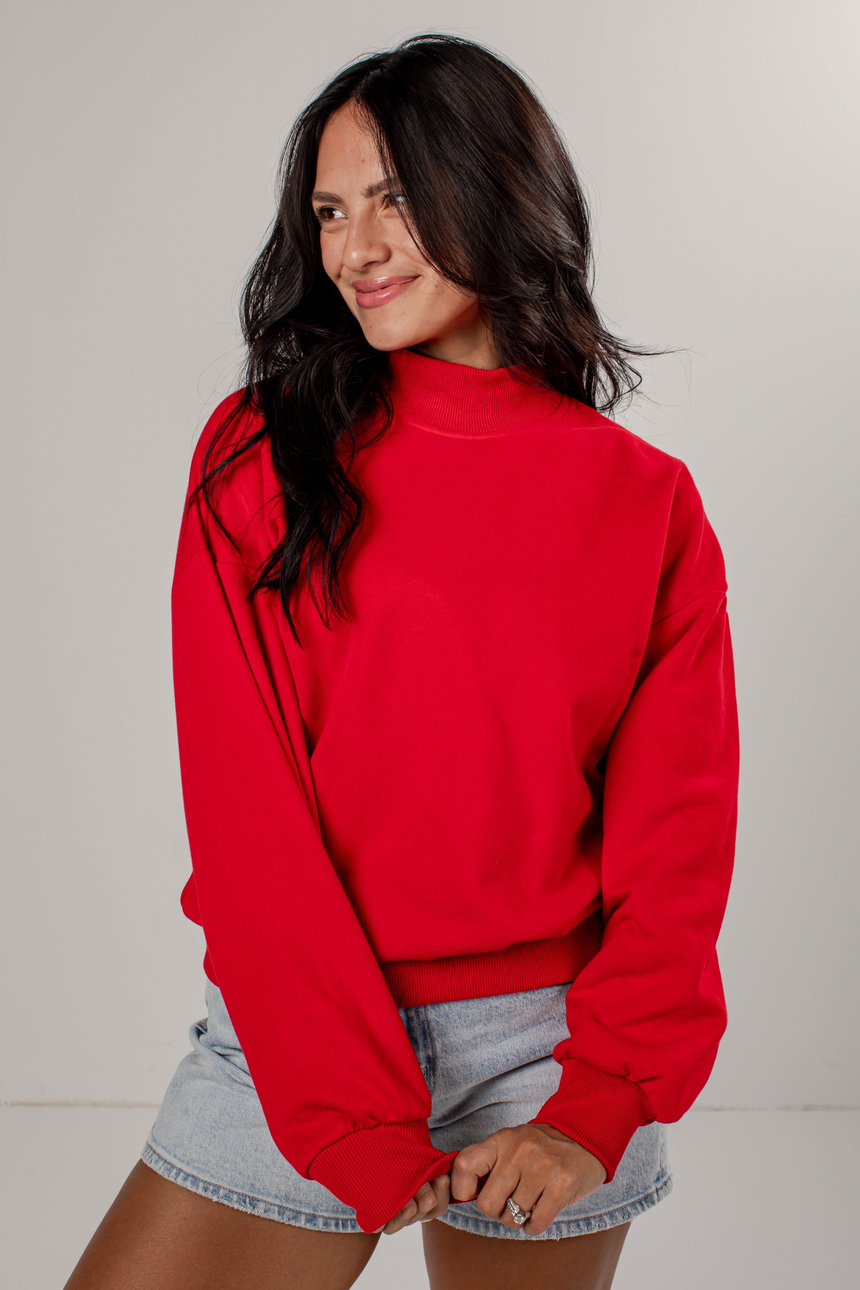 A woman wearing a bright red mock neck sweatshirt and light-wash denim shorts, posing against a neutral light gray studio background.