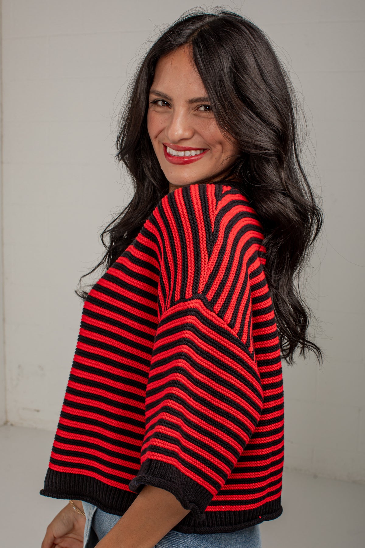 A woman with long dark hair is shown from the back and side, wearing a red and black striped knit sweater with wide three-quarter sleeves and a ribbed hem, against a plain light gray wall.