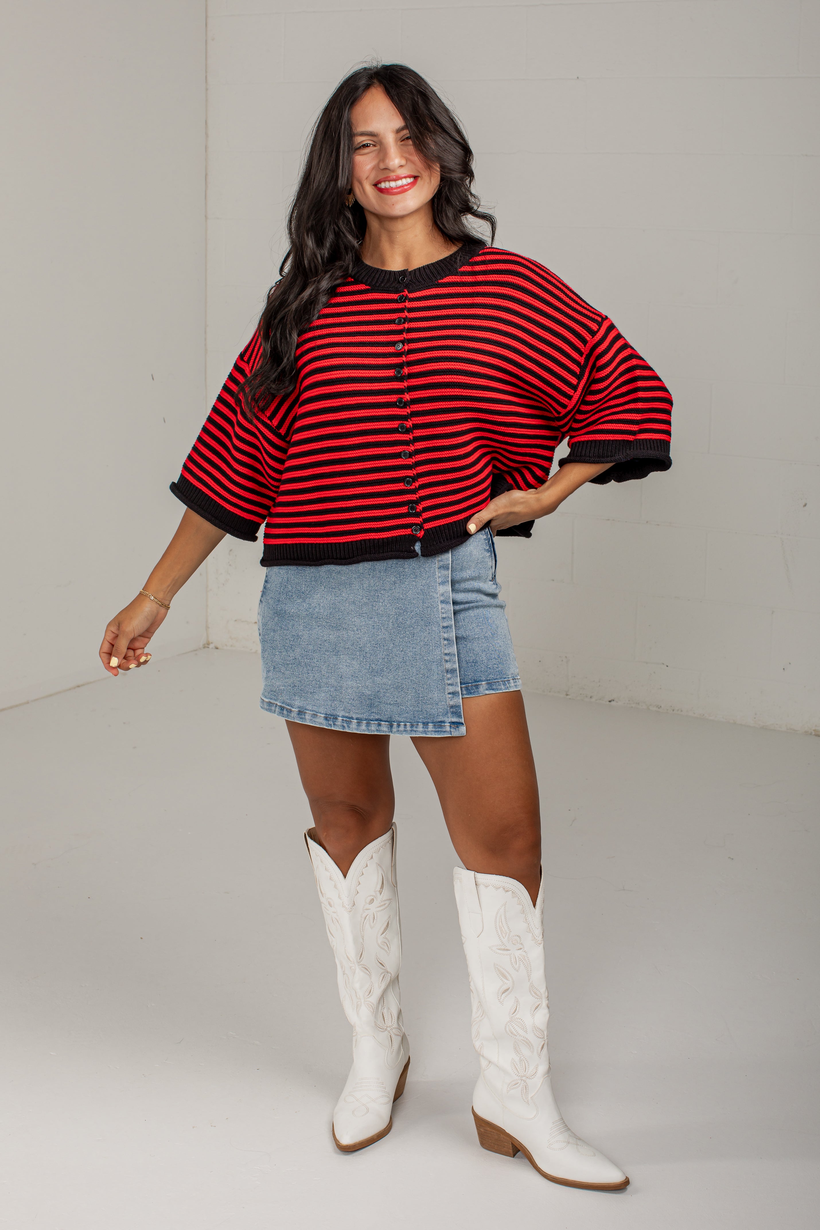A woman modeling a red and black striped knit poncho-style cardigan with a button front, paired with a light blue denim skirt and white cowboy boots, in a bright studio setting.