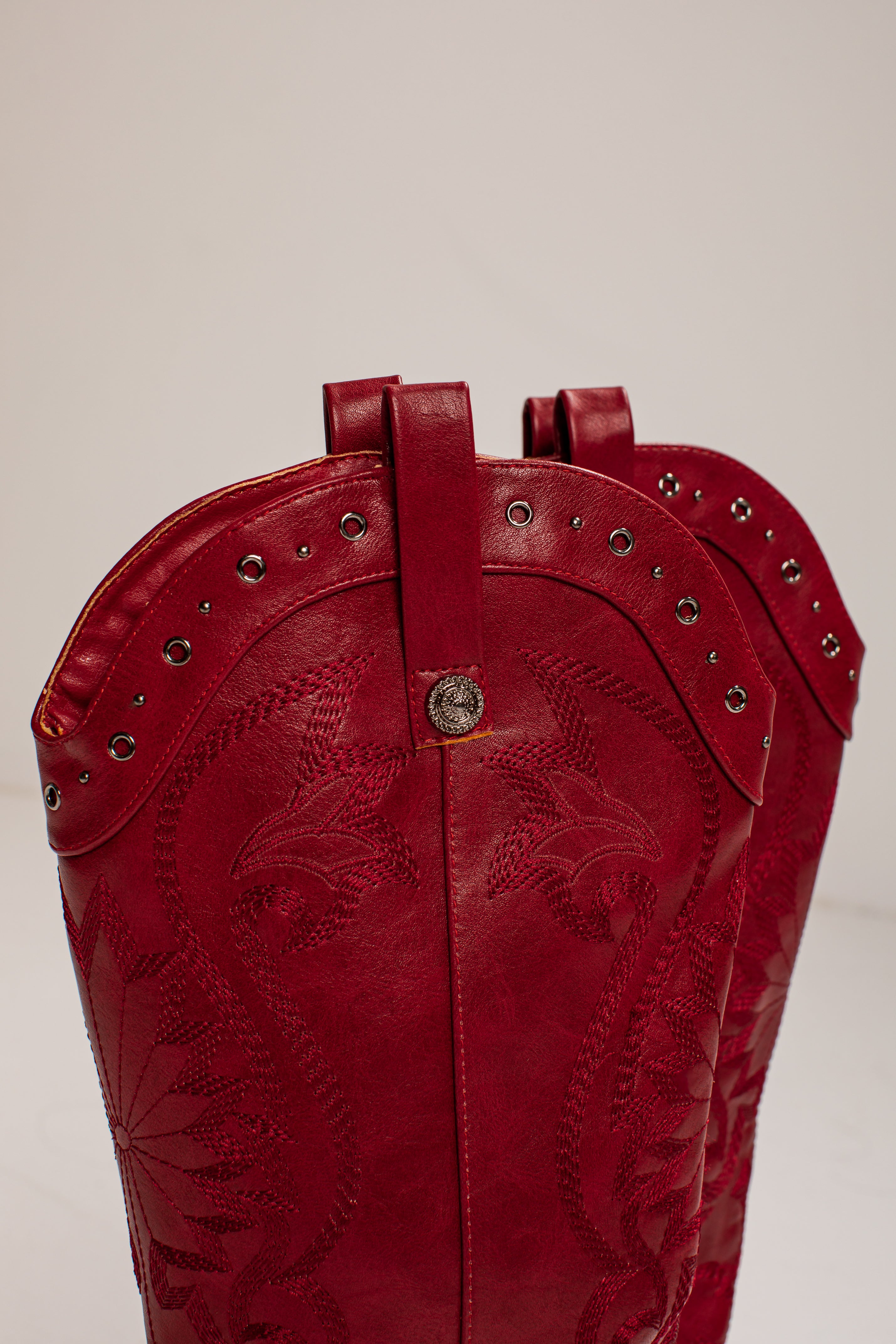 A close-up of a red leather cowboy boot showing the upper shaft with floral tooling, silver studs, and a central silver concho on a light gray background.