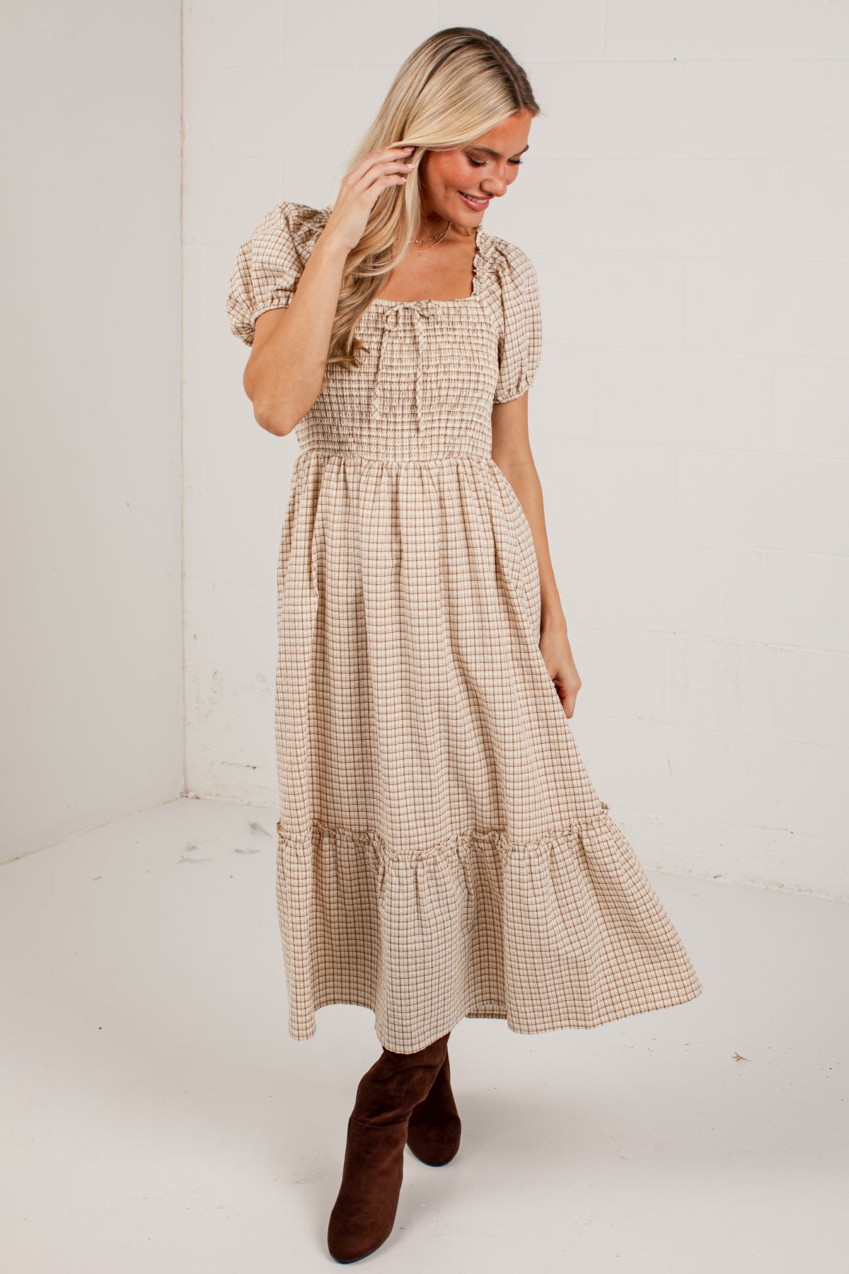 A woman wearing a beige and brown gingham midi dress with puff sleeves and a smocked bodice, paired with brown suede boots, standing in a white studio corner.
