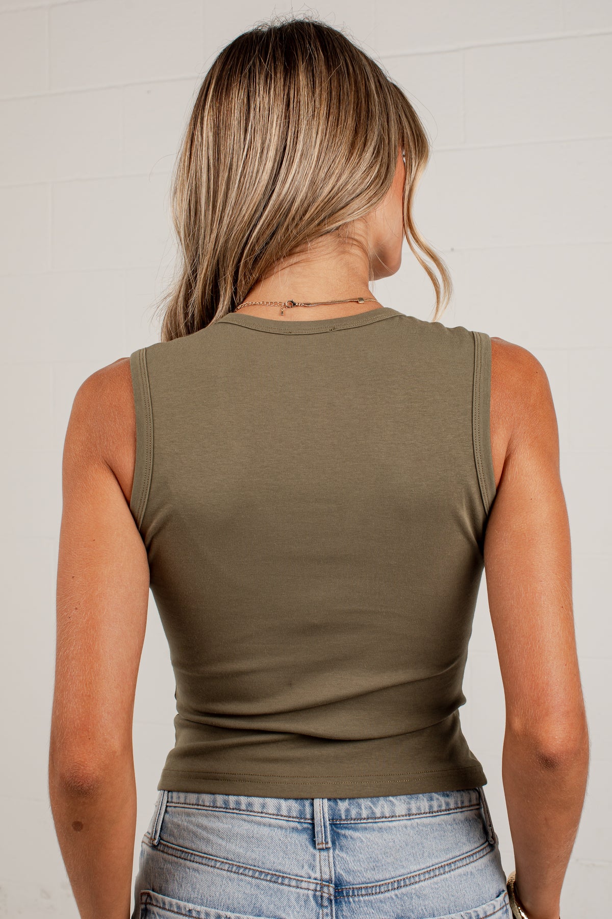 A woman viewed from behind wearing a ribbed sleeveless crop top in olive green and light-wash blue jeans, standing against a white paneled wall.