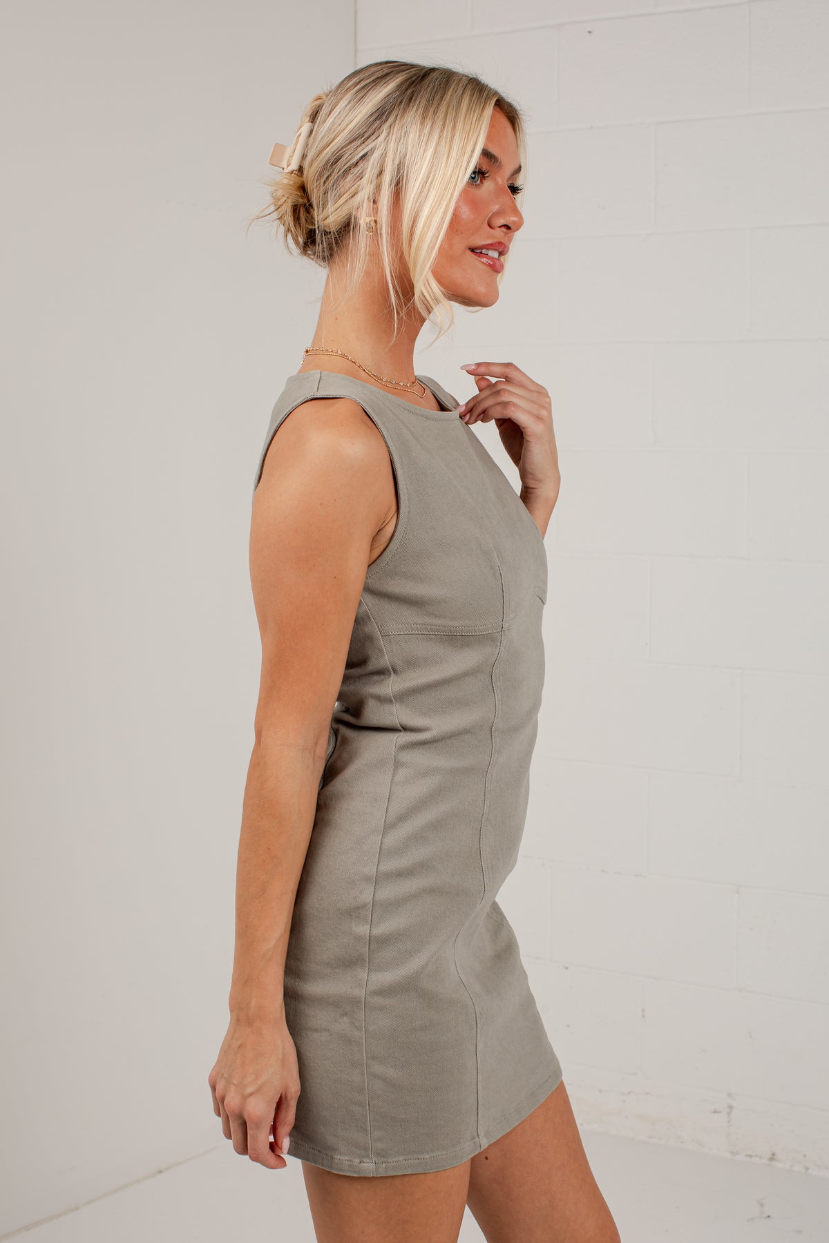 A blonde woman wearing a sleeveless taupe-gray mini dress, shown from a side profile against a white brick wall.
