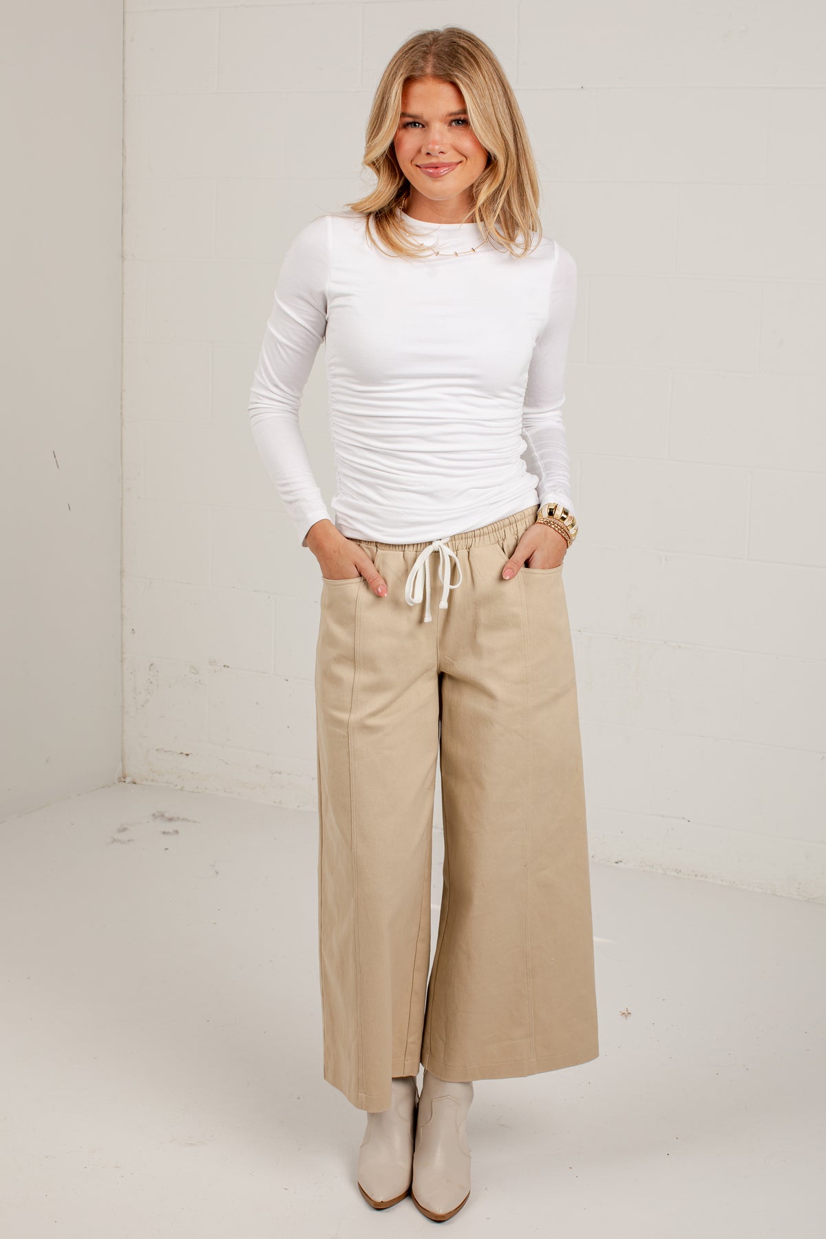 A woman modeling a white long-sleeve ruched top and beige wide-leg cropped pants with a drawstring waist, standing against a plain off-white studio wall.
