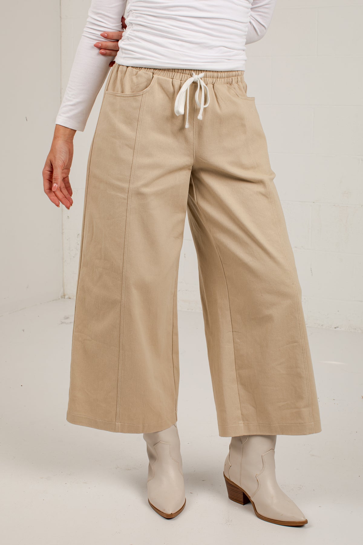 A person wearing beige wide-leg cropped pants with a white drawstring waist, paired with a white long-sleeve top and light beige ankle boots, photographed against a plain light wall.