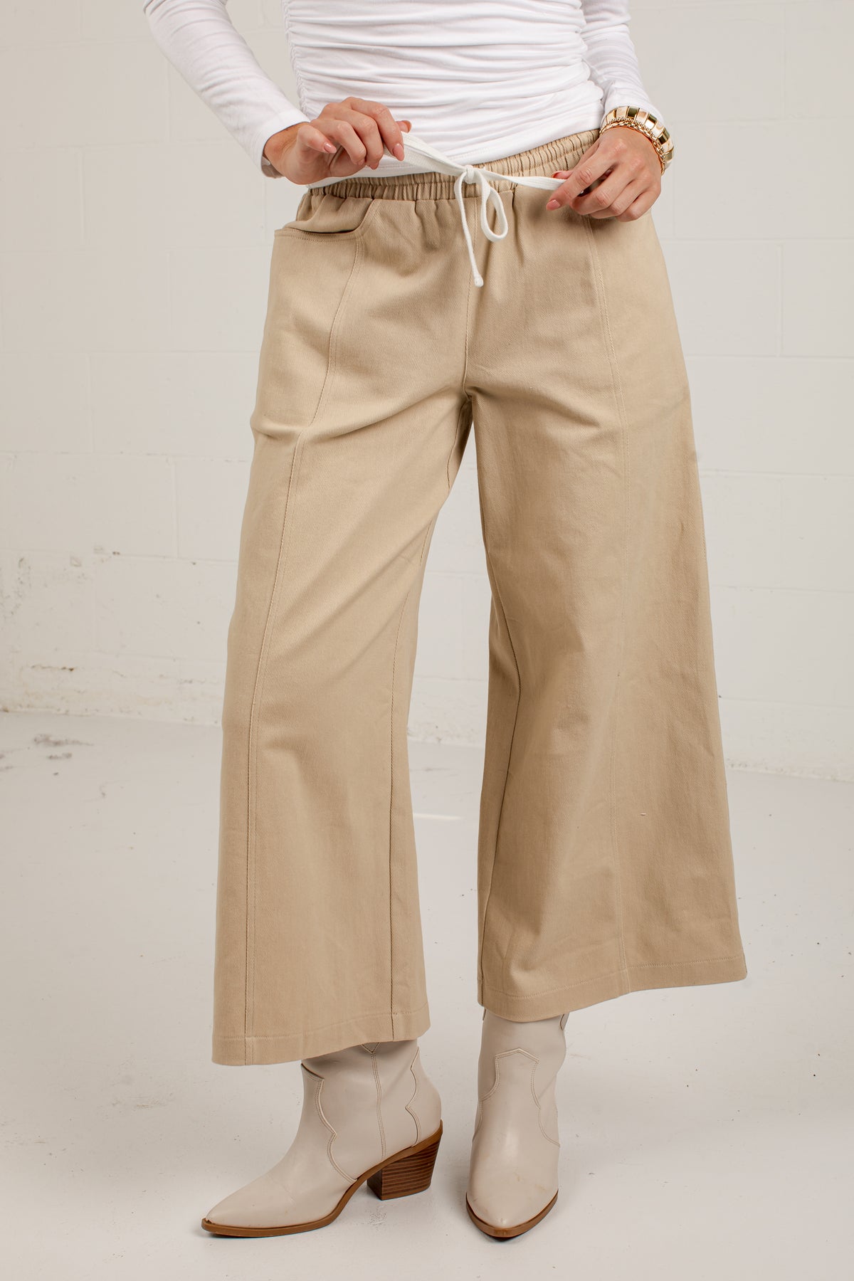 A person wearing beige wide-leg cropped pants with an elastic drawstring waist, paired with a white long-sleeve top and light beige heeled boots, standing against a light off-white wall.