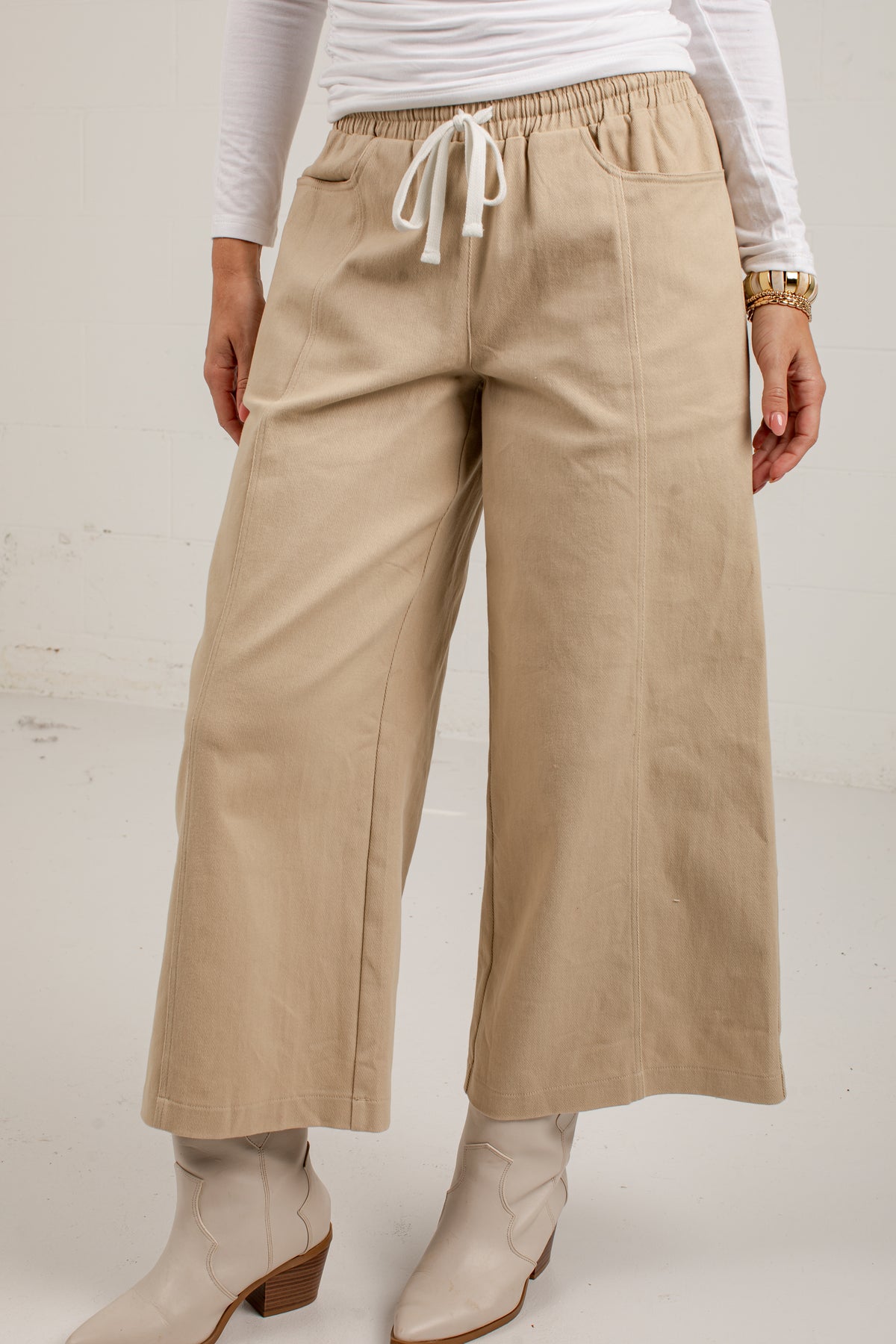A person wearing beige wide-leg cropped pants with a white drawstring waist, paired with a white long-sleeve top and light-colored heeled boots, photographed against a plain light background.