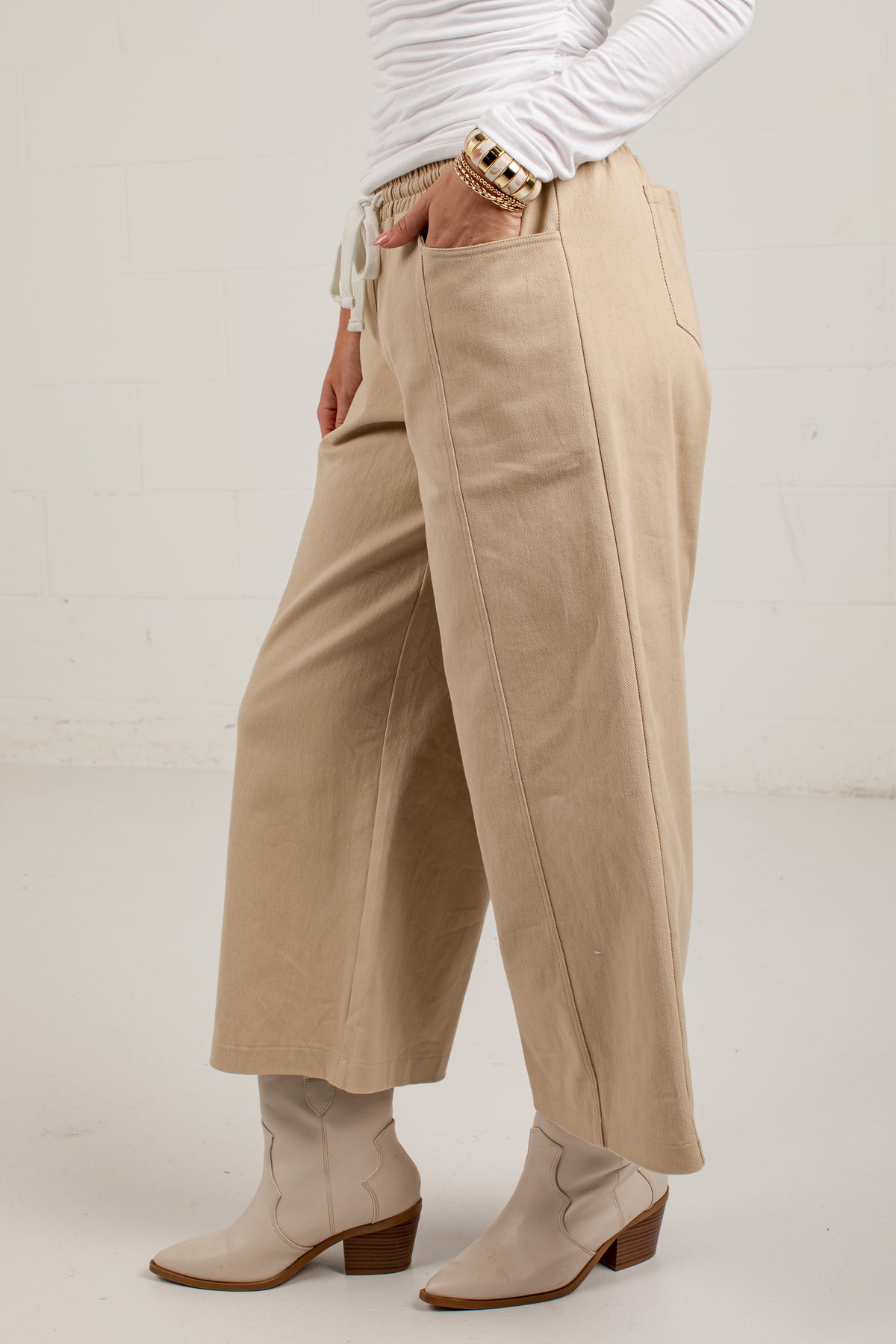 A person wearing wide-leg cropped pants in a light beige or tan color, styled with a white long-sleeve top and beige heeled ankle boots against a plain off-white studio background.