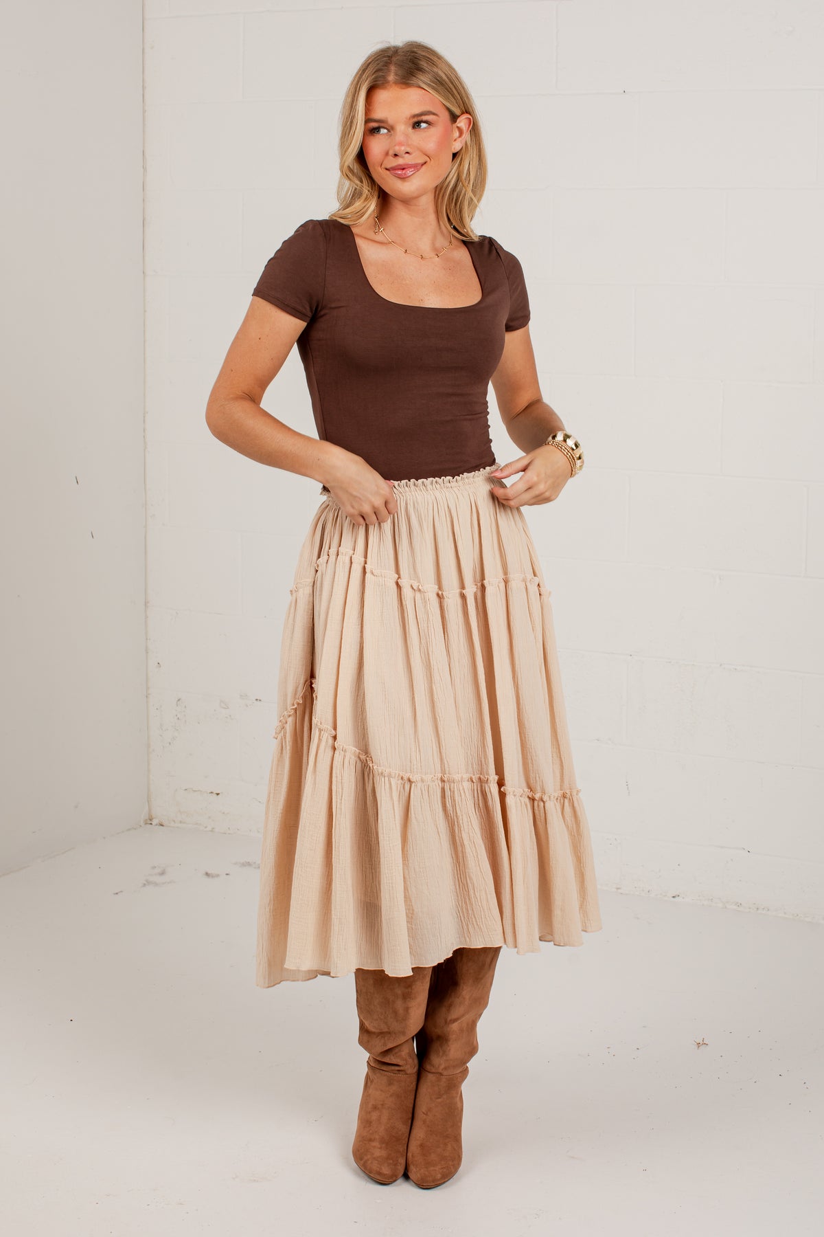 A woman modeling a tiered midi skirt in a light beige color, paired with a brown short-sleeve top and brown boots, standing against a plain white studio wall.