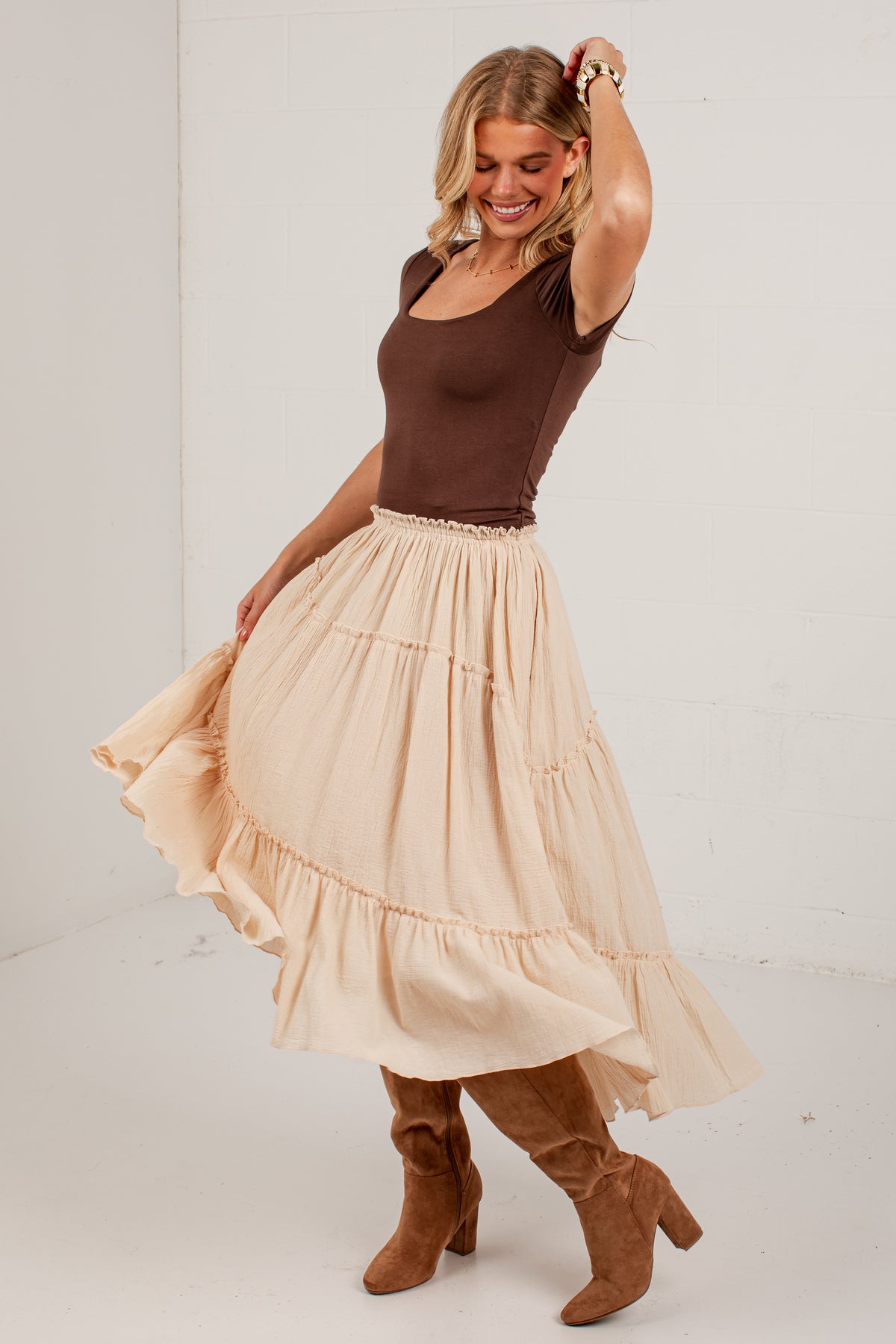 A woman modeling a tiered beige maxi skirt with a ruffled hem, paired with a brown scoop-neck top and brown suede knee-high boots, set against a plain white studio background.