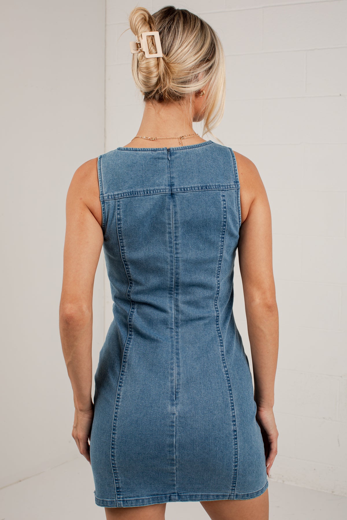 A woman viewed from behind wearing a sleeveless blue denim dress, standing against a light-colored brick wall.