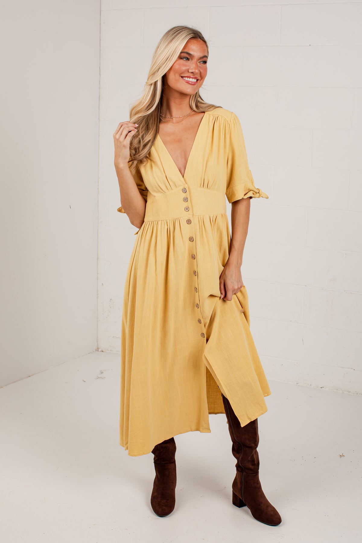 A woman modeling a mustard yellow midi dress with a button-down front, ruffled short sleeves, and an asymmetrical hem, paired with brown knee-high boots in a bright studio setting.