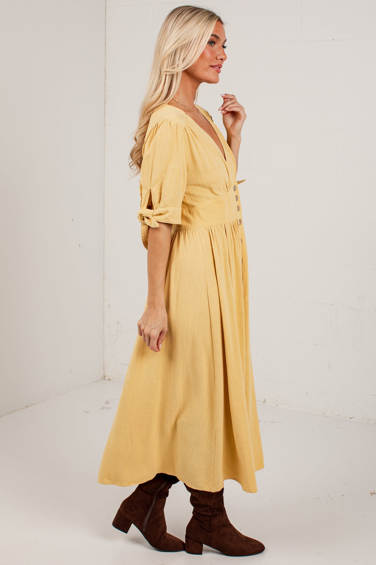 A woman modeling a mustard yellow midi dress with puff sleeves, a V-neckline, and a gathered waist, paired with brown suede ankle boots, set against a white studio background.