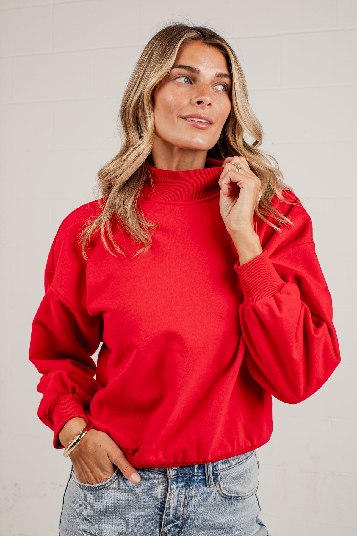 A woman wearing a red mockneck sweatshirt and light blue jeans, standing against a solid light gray background.