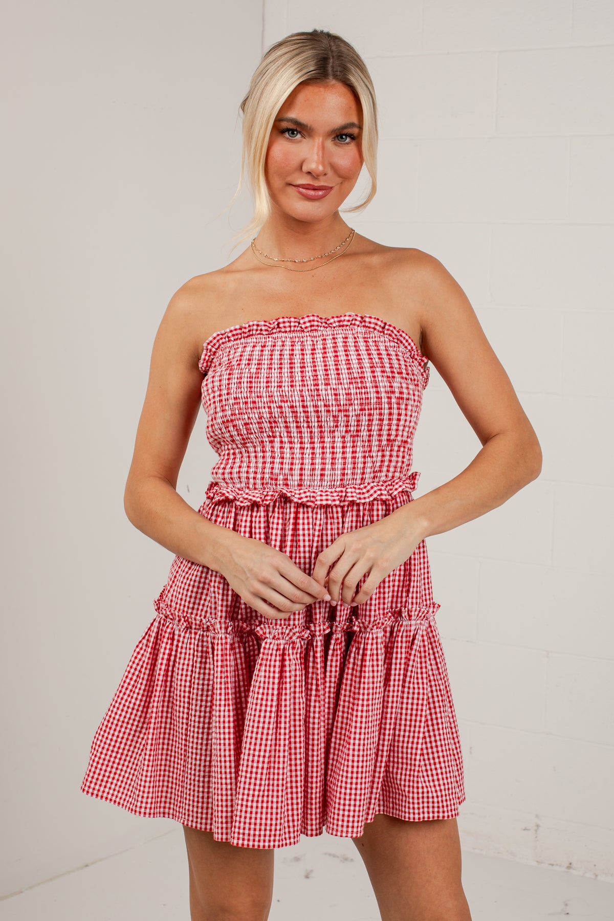 A woman modeling a strapless red and white gingham mini dress with a smocked bodice and ruffled tiered skirt, standing against a light off-white brick wall.