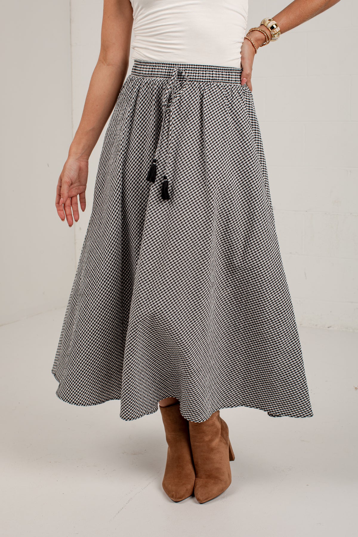 A woman wearing a black and white micro-checkered midi skirt with a drawstring waist, styled with a white top and brown suede ankle boots.
