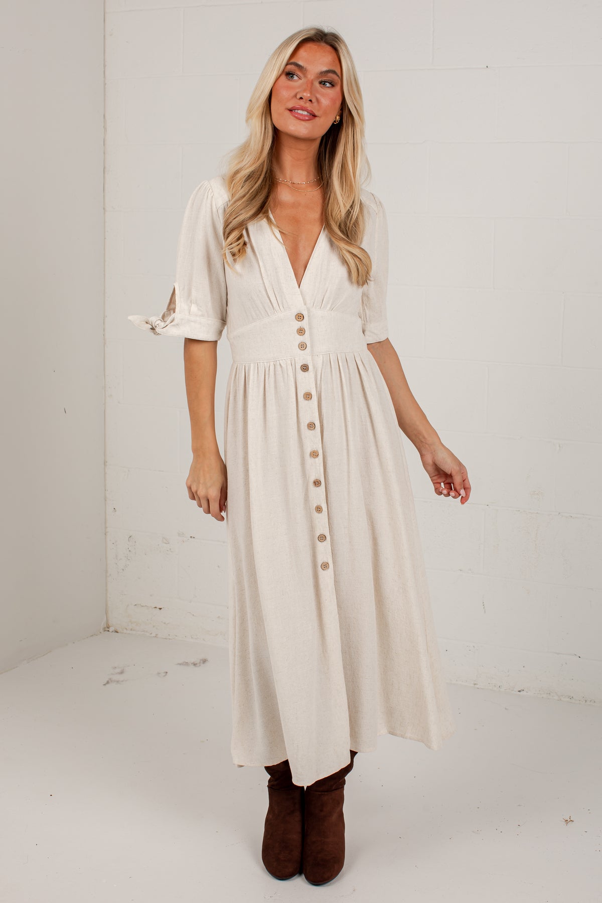 A woman modeling a cream-colored midi dress with a button-down front, short sleeves with tie details, and a gathered waist, paired with brown boots in a white studio setting.