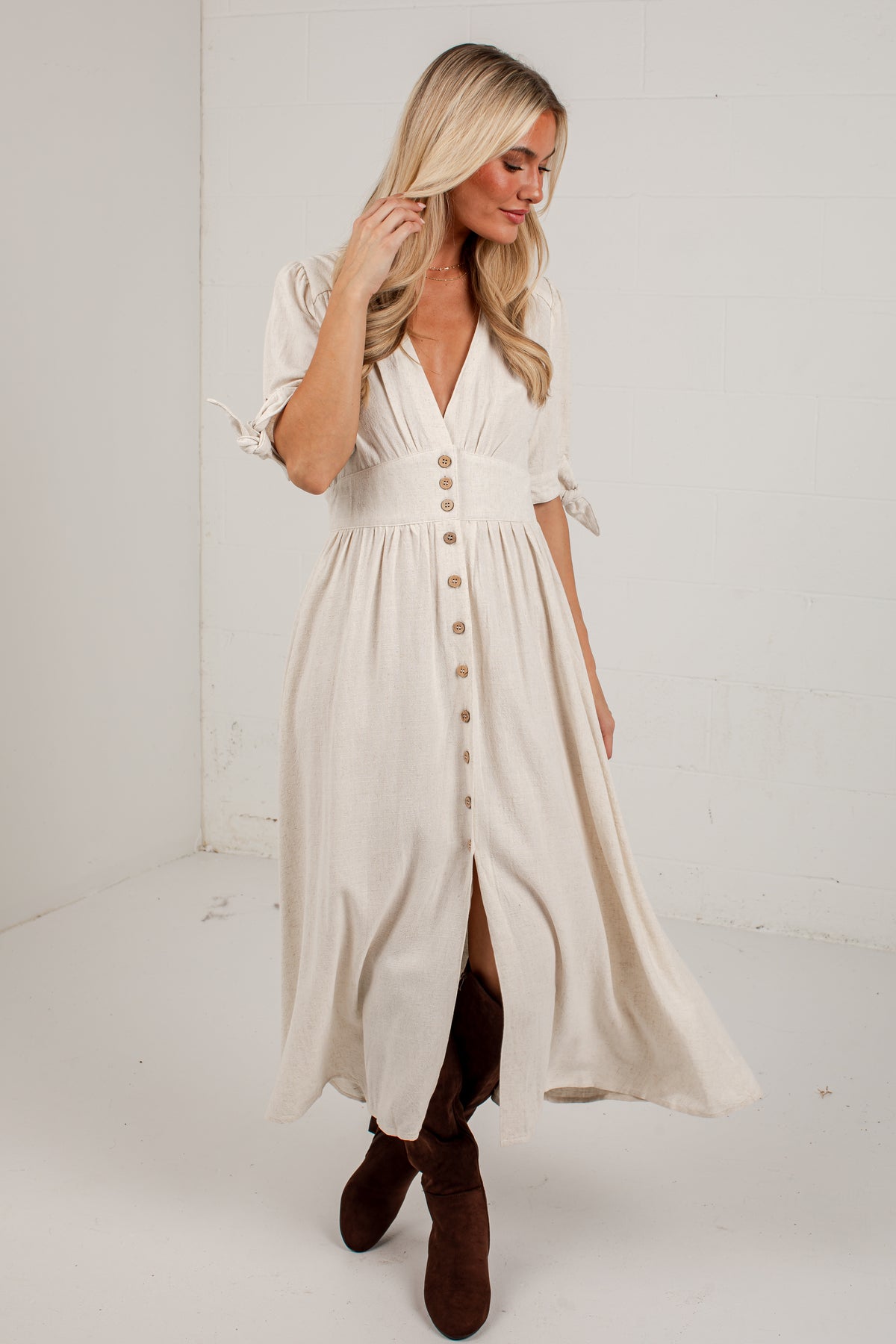 A woman modeling a cream-colored midi dress with a button-down front, ruffled sleeves, and a high slit, paired with brown boots in a bright studio setting.