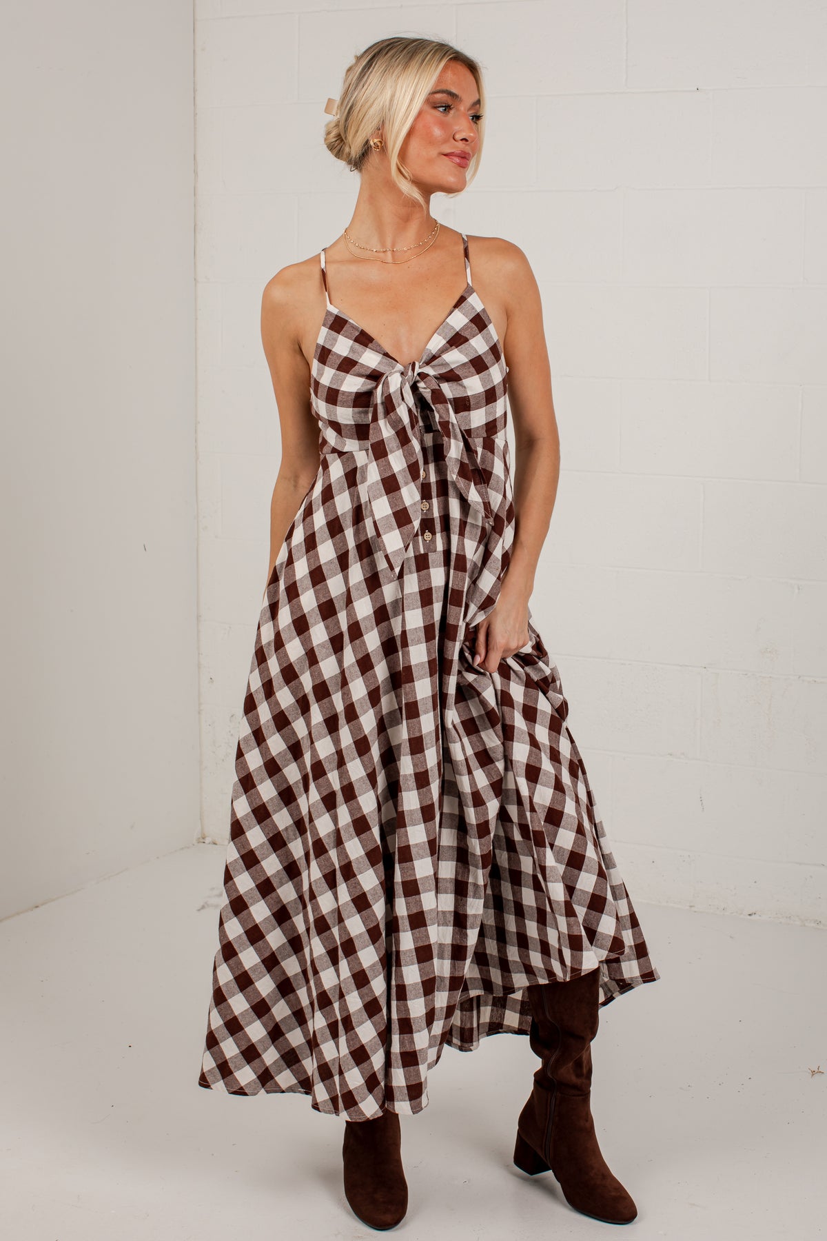 A woman modeling a brown and beige gingham maxi dress with spaghetti straps, a sweetheart neckline, and a front tie detail, paired with brown suede boots, set against a white brick wall background.
