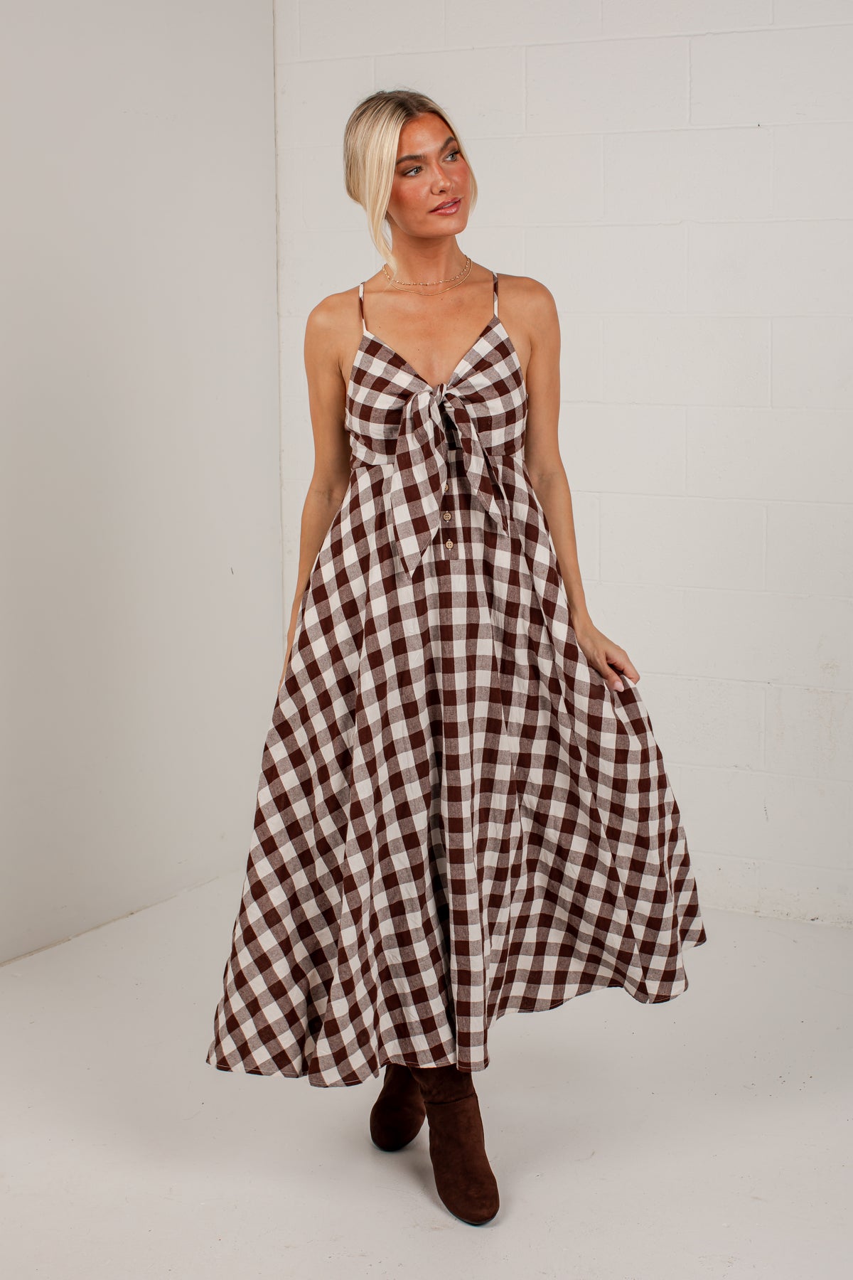 A woman modeling a brown and beige gingham maxi dress with spaghetti straps, a sweetheart neckline, and a large front bow, paired with brown suede boots, set against a white studio background.