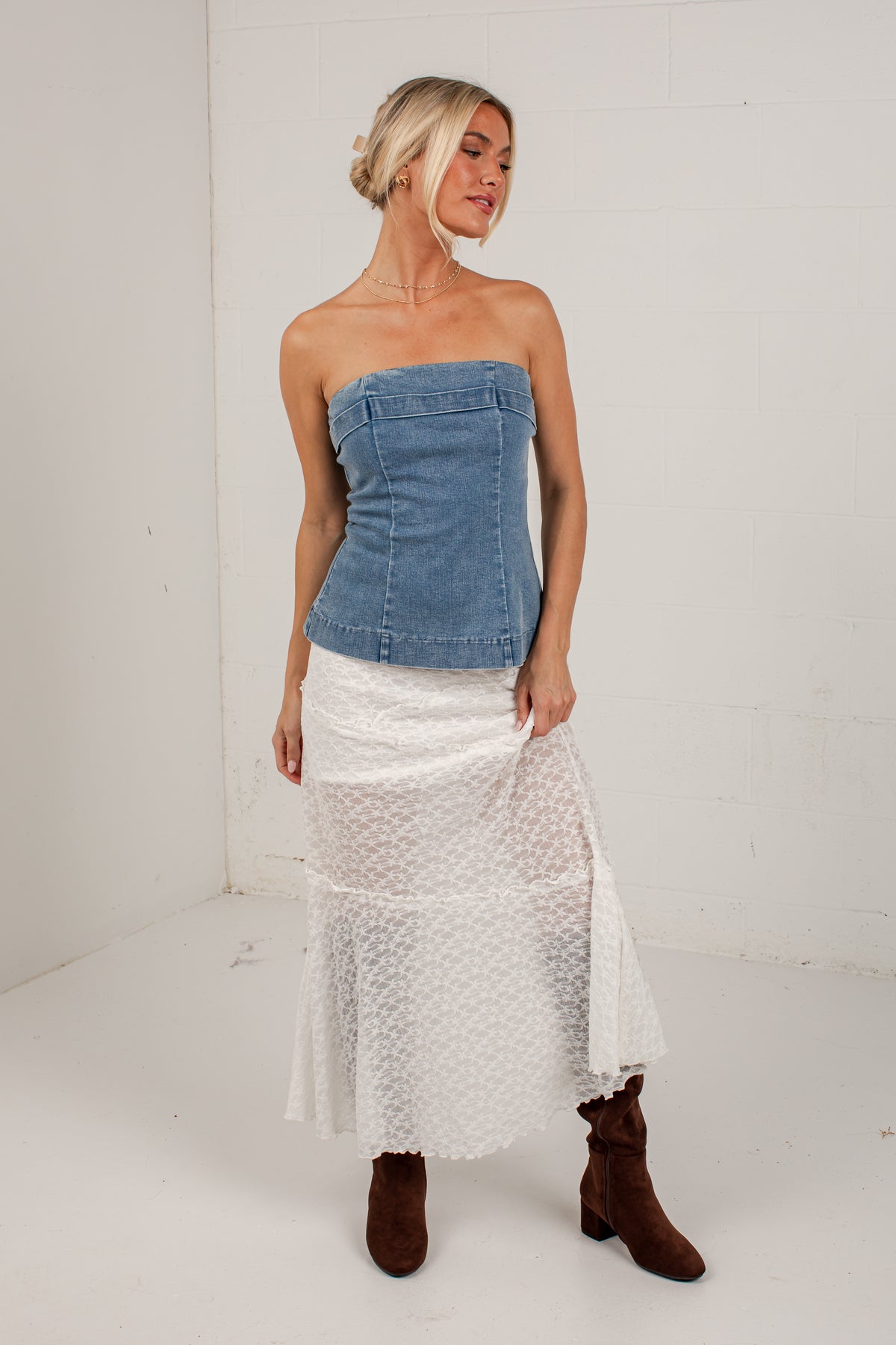 A woman modeling a strapless blue denim corset-style top paired with a white textured midi skirt and brown boots, standing against a white studio wall.