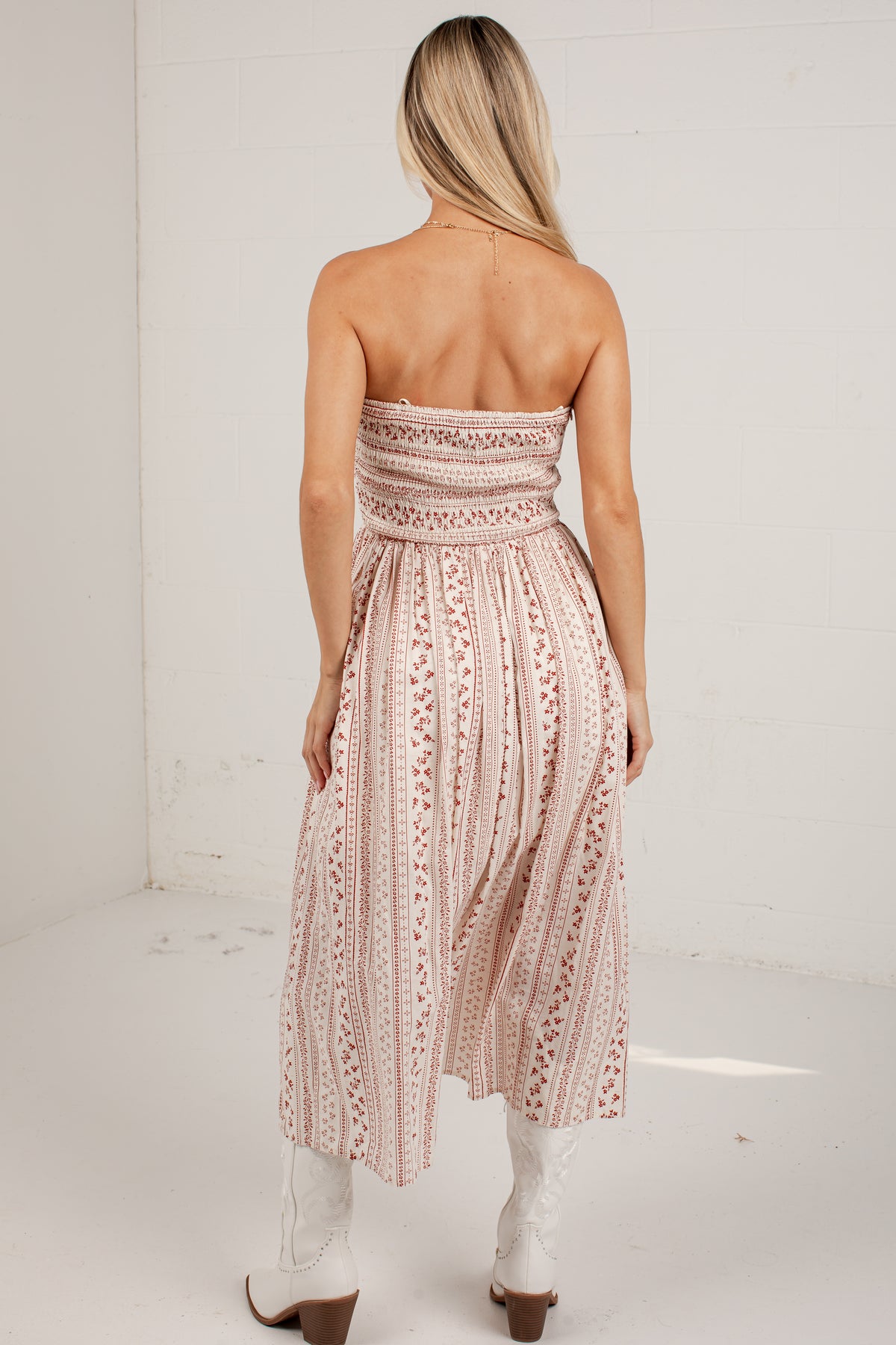 A woman shown from the back wearing a strapless midi dress with a smocked bodice and a pleated skirt, featuring a vertical floral pattern in dusty pink and white. She is wearing white cowboy boots in a bright, white-walled room.