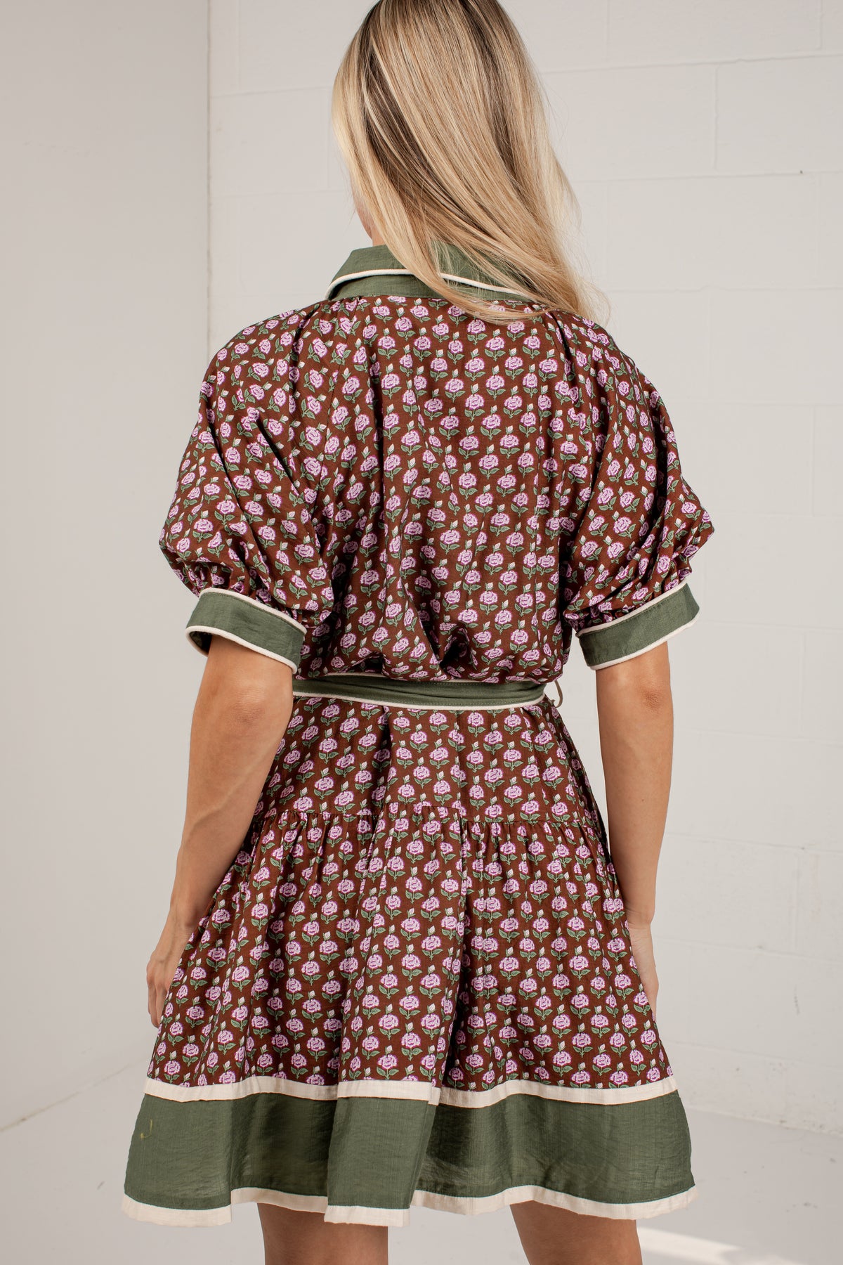 A woman viewed from behind wearing a short puff-sleeve dress with an allover floral print in brown, pink, and green, featuring a matching green waist tie and a green hem with cream piping.