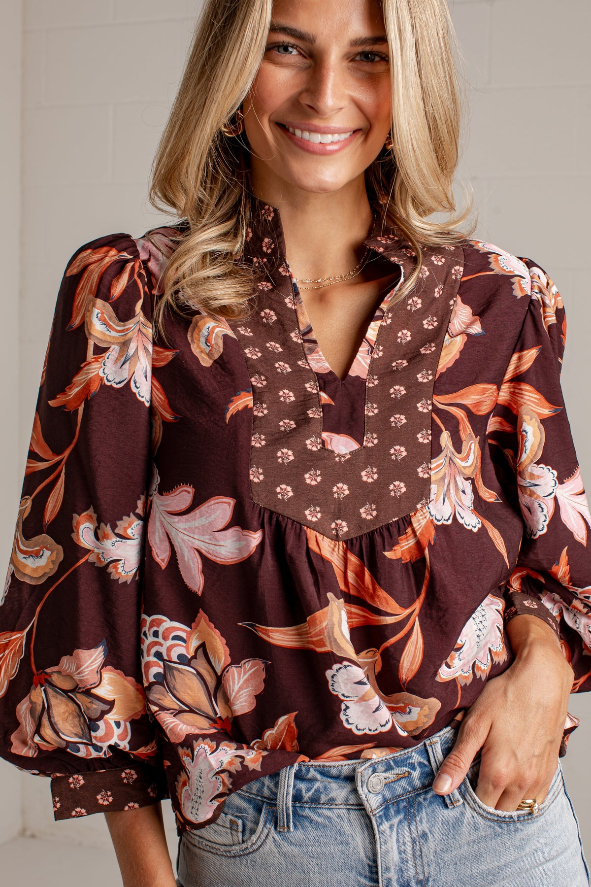A woman wearing a brown floral blouse with puff sleeves and a V-neckline, paired with light-wash blue jeans.